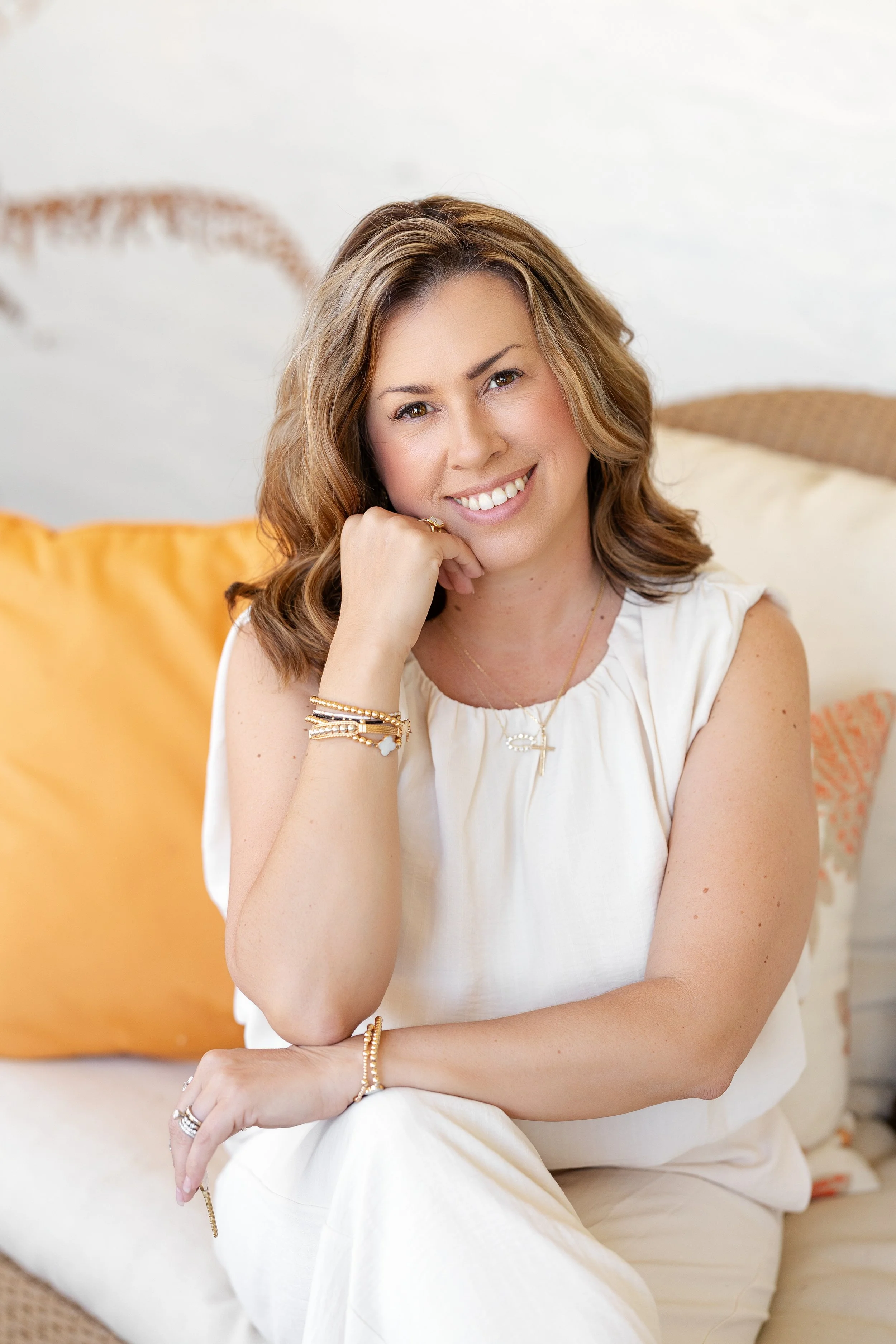 A woman with wavy brown hair and a warm smile sitting on a beige sofa with colorful cushions, wearing a white sleeveless top and jewelry, in a bright room with a white wall background.