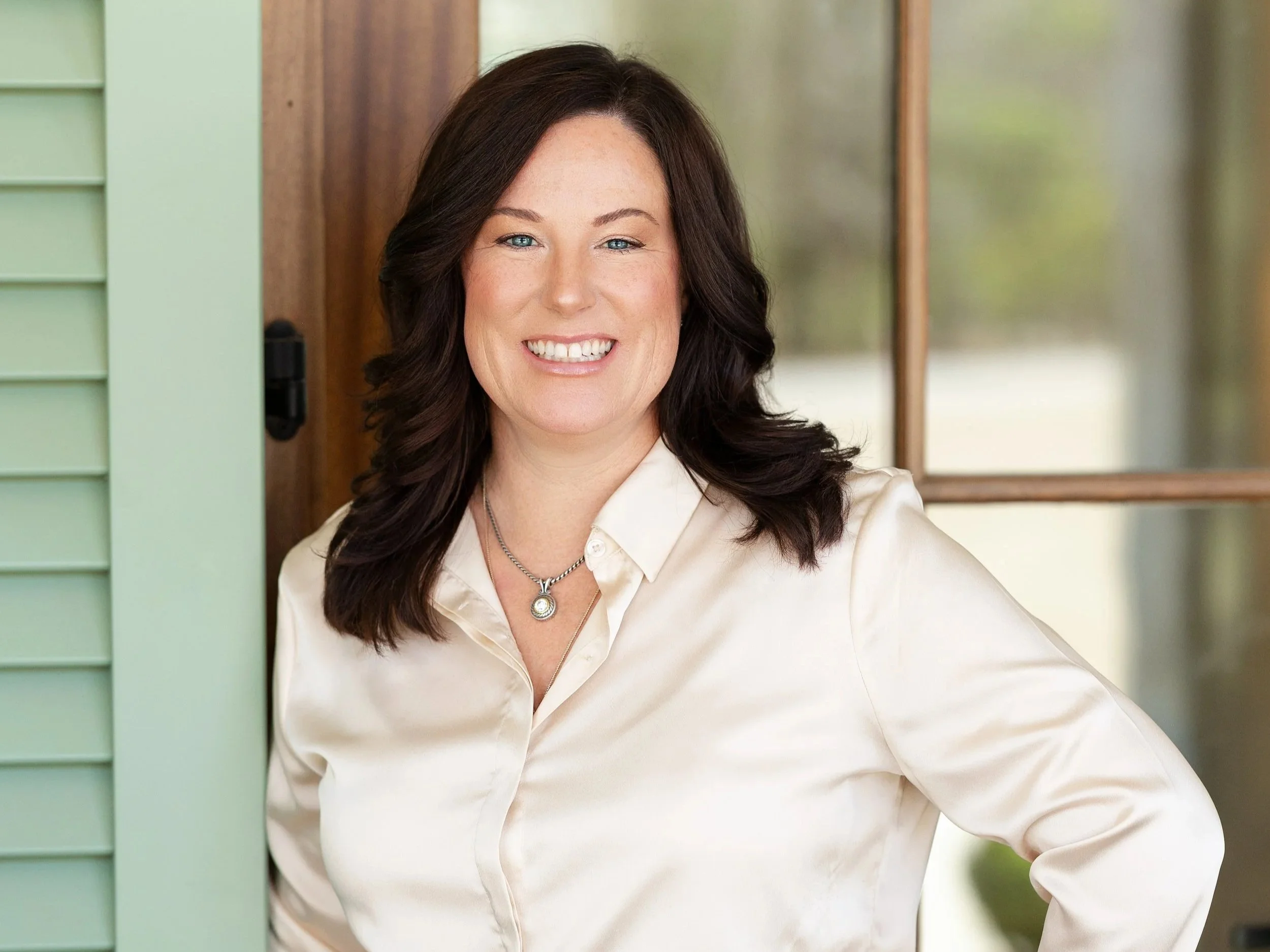 A woman with shoulder-length dark brown hair, blue eyes, and a wide smile showing teeth, wearing a beige satin blouse and a silver necklace with a round pendant, standing indoors in front of a wooden wall and a window.