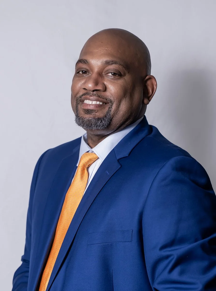 A bald African American man with a beard, wearing a blue suit and a yellow tie, smiling confidently at the camera against a plain light background.