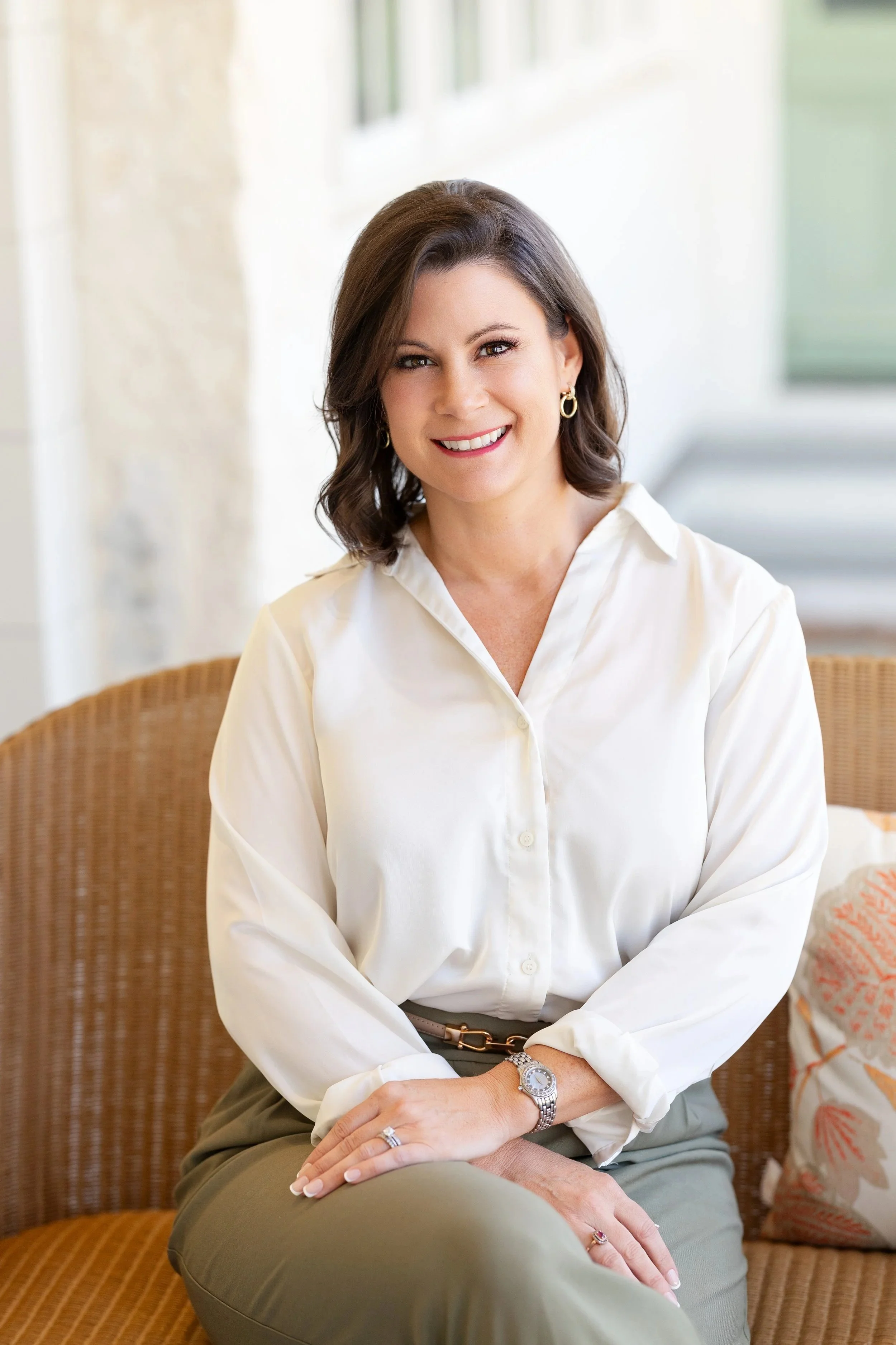 A woman with short brown hair, wearing a white button-up shirt, a wristwatch, and jewelry, sitting on a wicker chair in a bright, airy room with white walls and a window.
