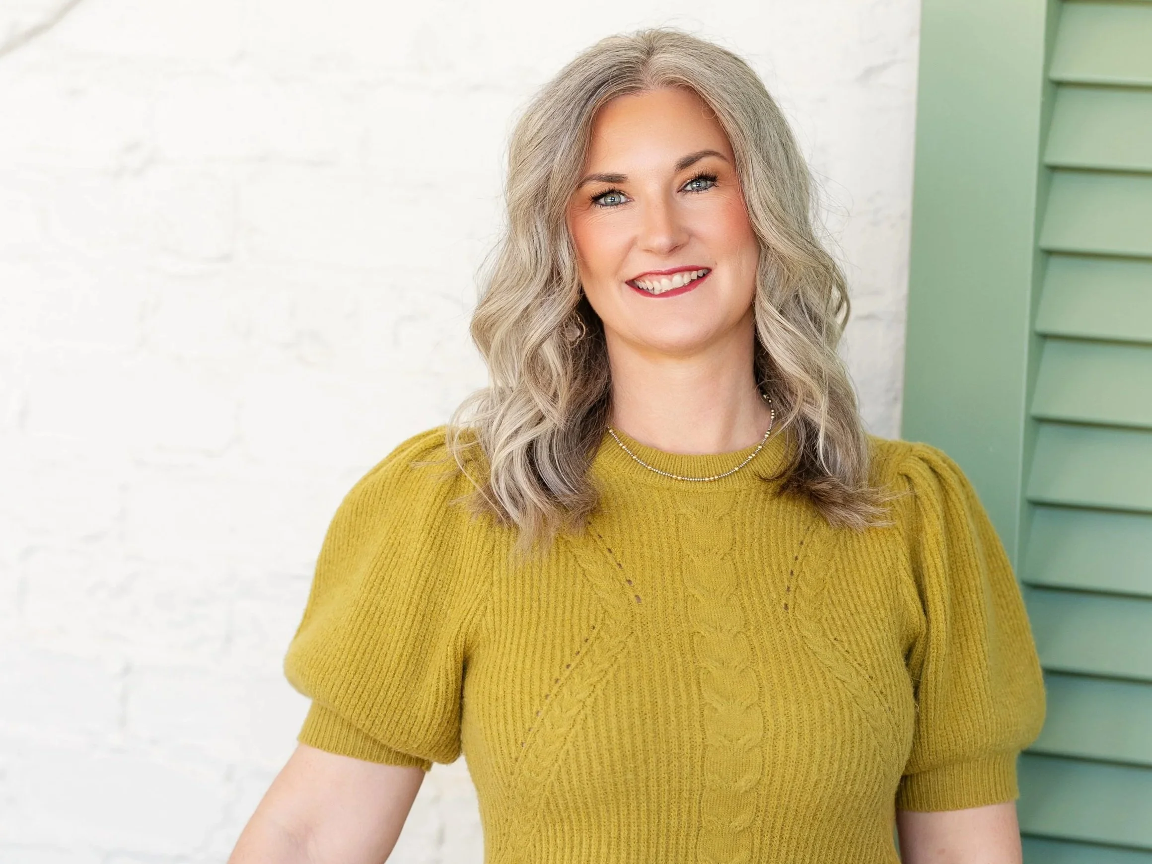 A woman with long wavy gray hair and fair skin smiling, wearing a mustard yellow knit sweater and a beaded necklace, standing against a white brick wall with a green cabinet on her right.