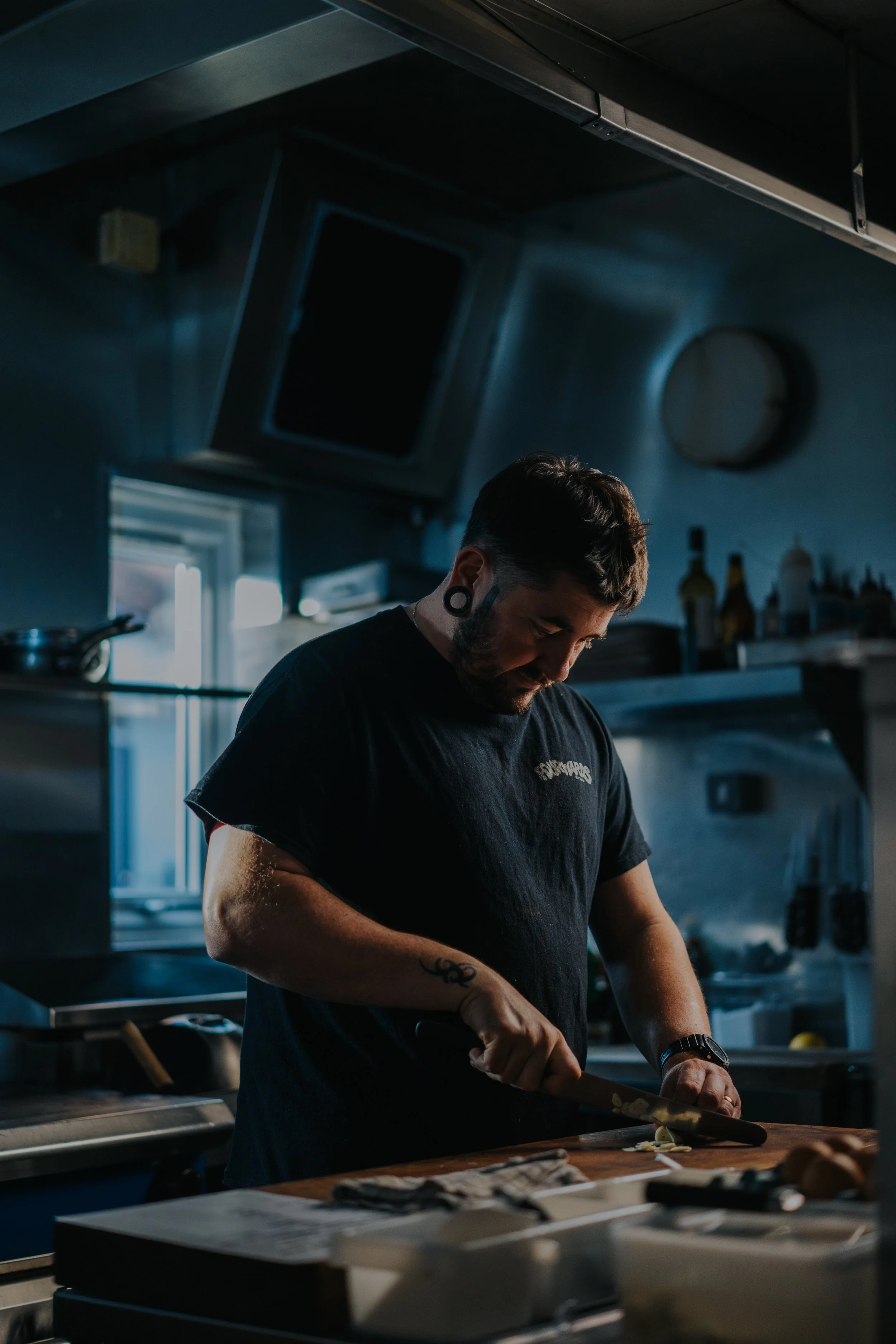 A man with tattoos on his arm and wearing a black T-shirt is chopping ingredients on a wooden cutting board in a dimly lit kitchen.