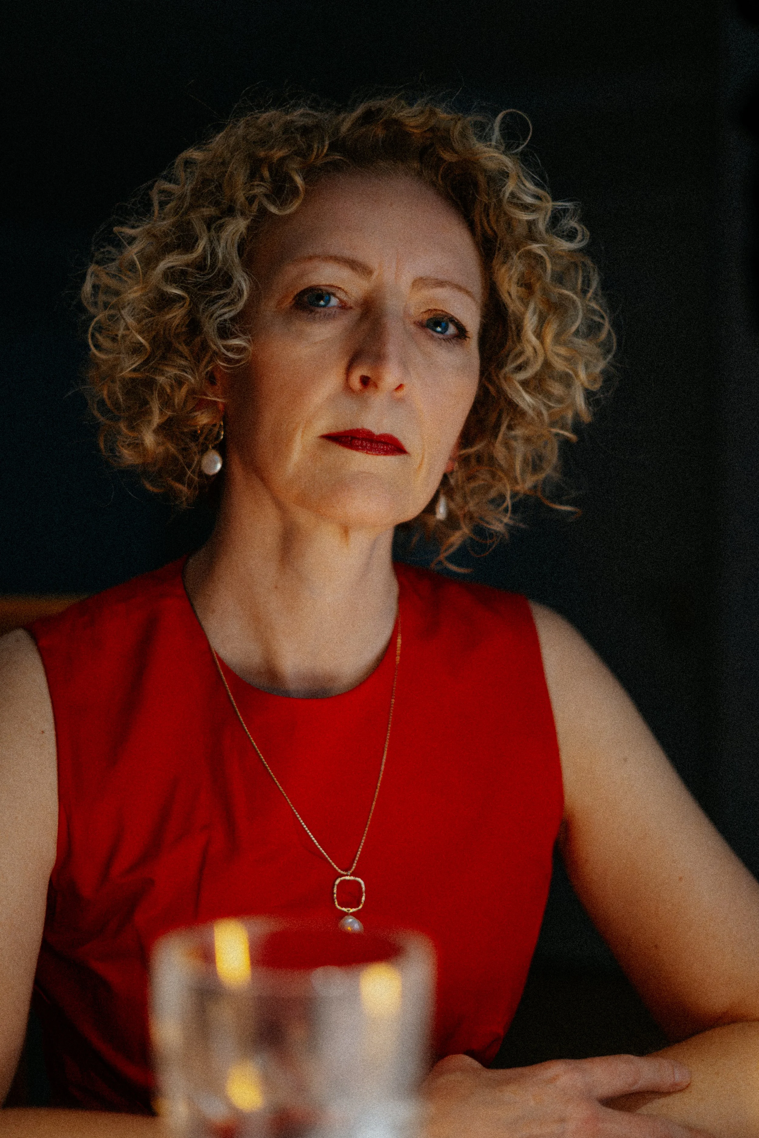 A woman with curly blonde hair, wearing a sleeveless red top, sitting at a table with a glass in the foreground. She has pearl earrings and a gold necklace with a pendant. She has a serious expression and is looking directly at the camera. The backgr