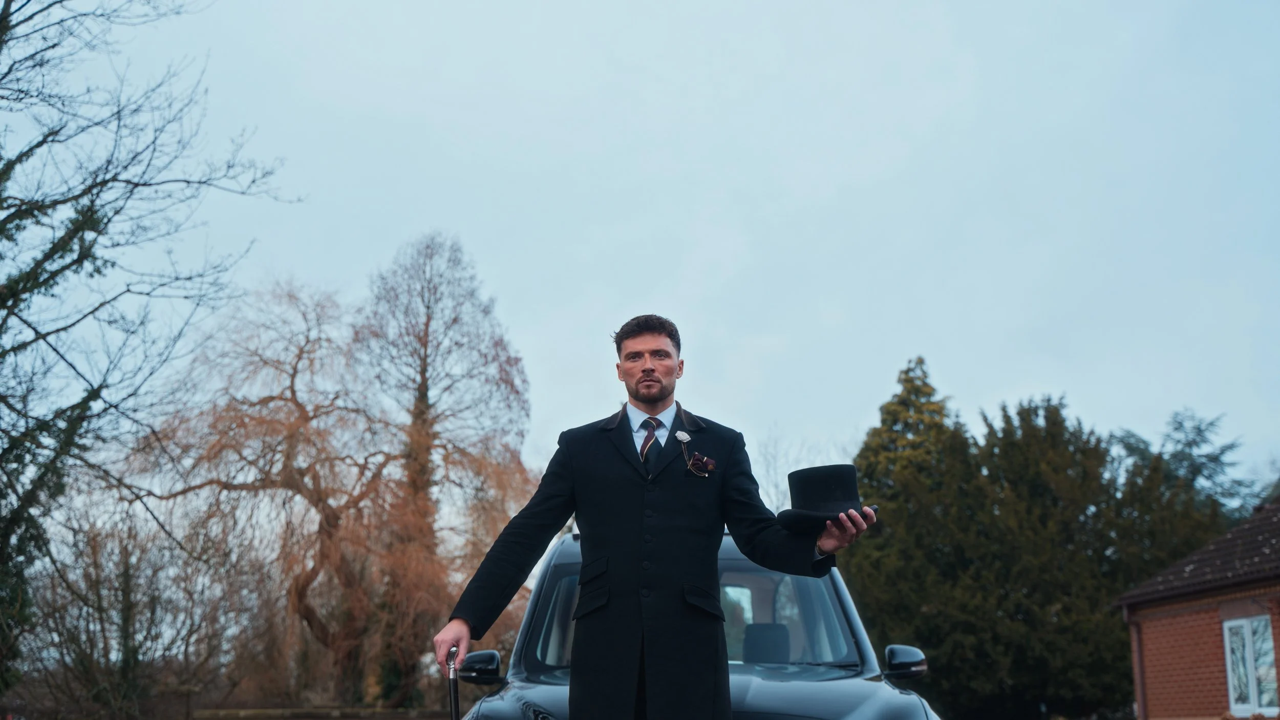 A man dressed in vintage formal attire standing in front of a black car, holding a top hat in one hand and a cane in the other, with Trees and a house in the background.