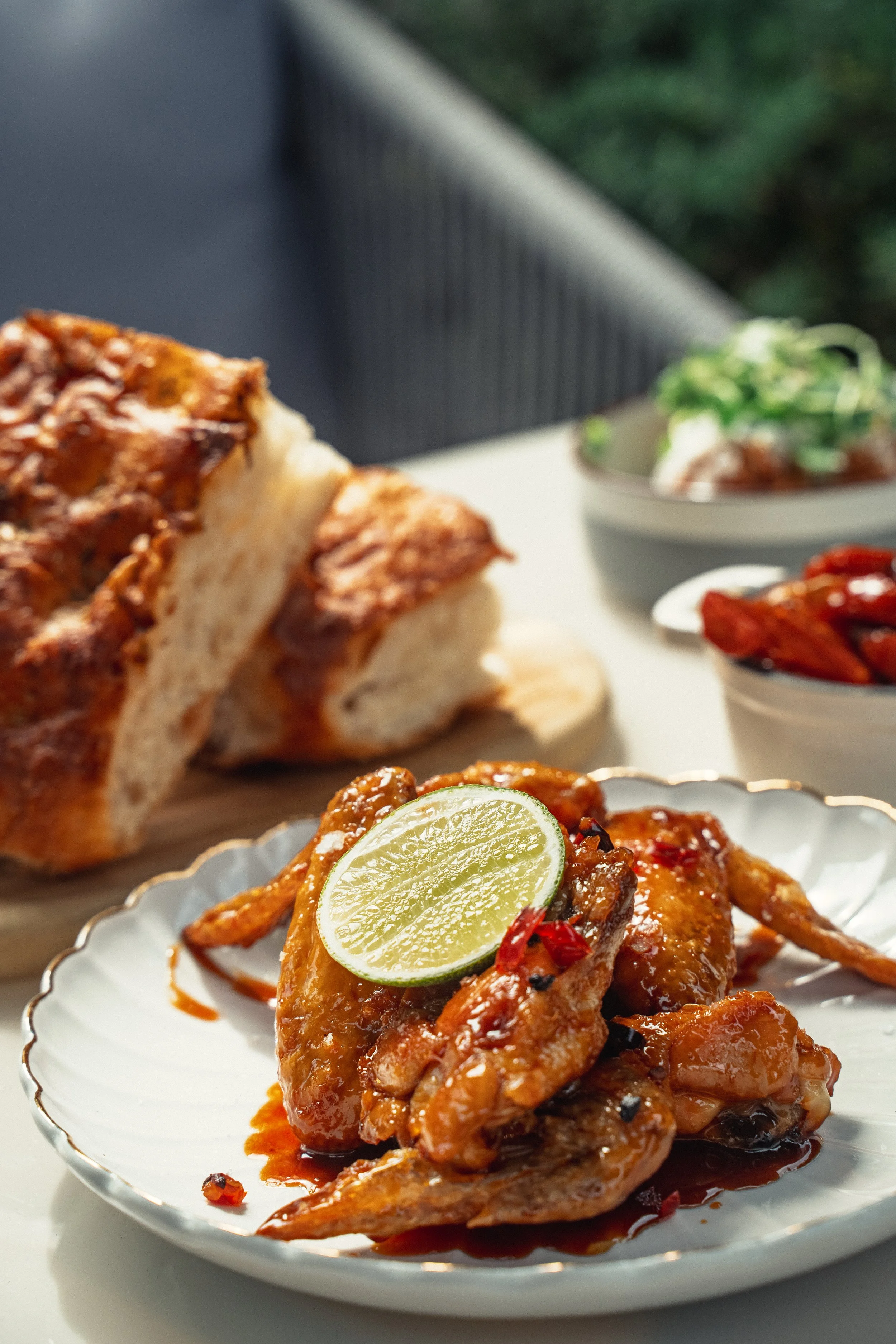 Close-up of spicy chicken wings with lime on a white plate, with bread and small bowls of different side dishes in the background.