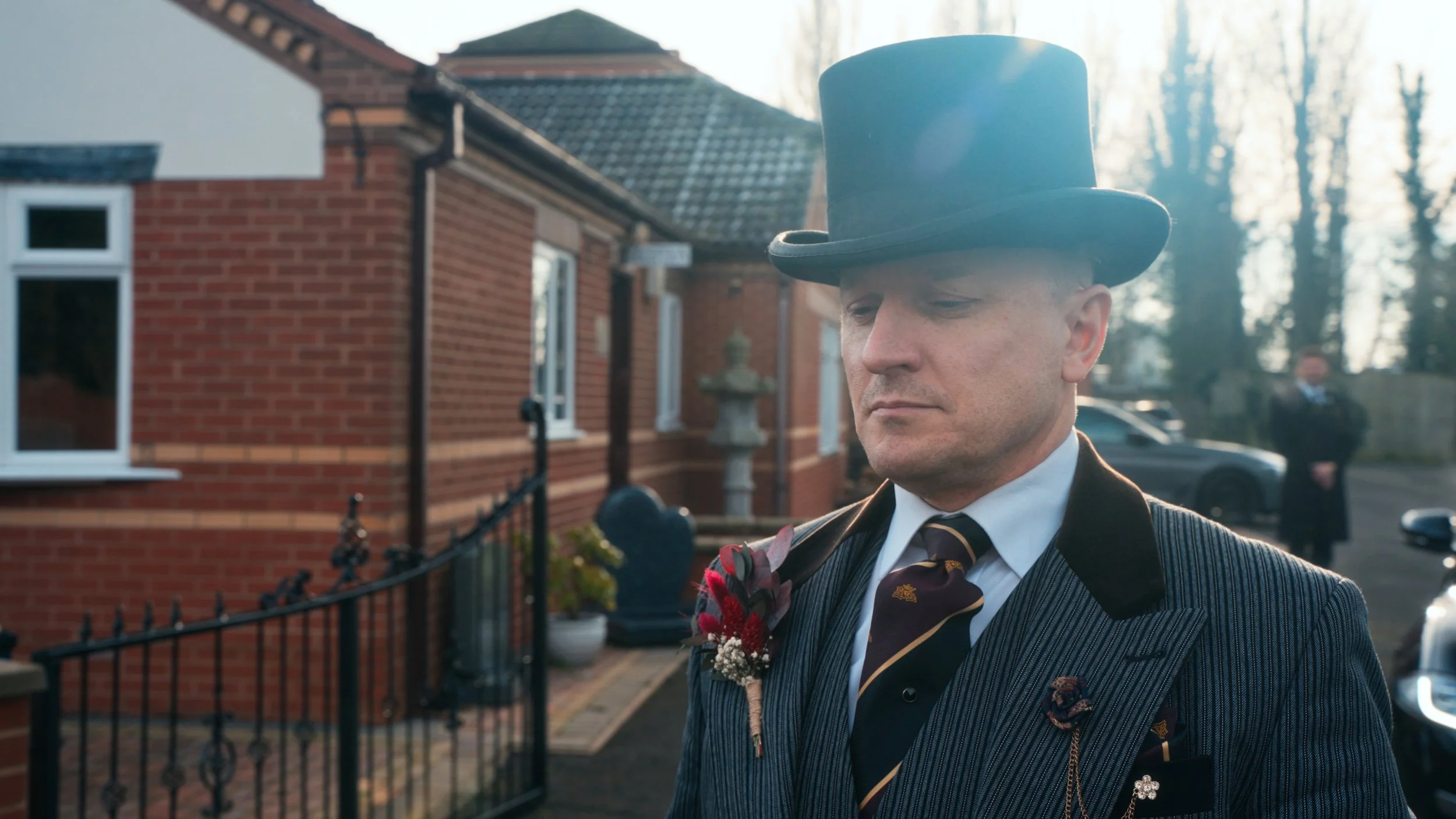 A man in a vintage style suit and tie, wearing a tall black top hat with a solemn expression, standing outdoors in front of a brick house with a garden and another man in the background.