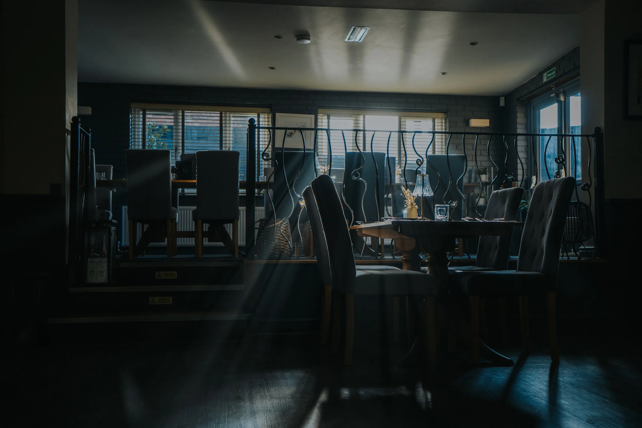 A dimly lit restaurant or cafe interior with a raised dining area and a round table with chairs, decorative items, and large windows in the background.