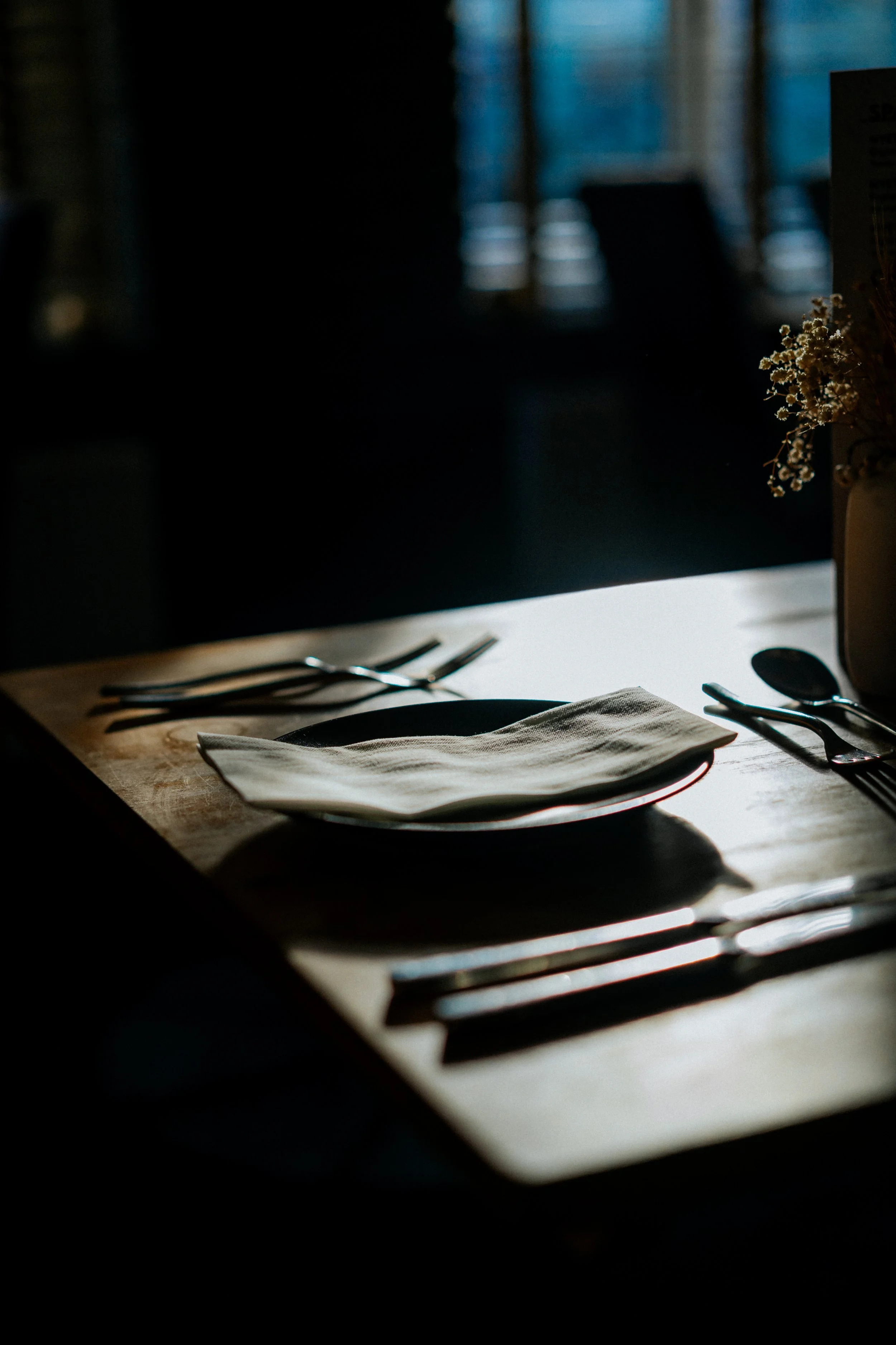 A dining table set with a white napkin on a black plate, silverware including forks, knives, and spoons, and a vase with white flowers, in a dimly lit room with natural light coming from a window in the background.