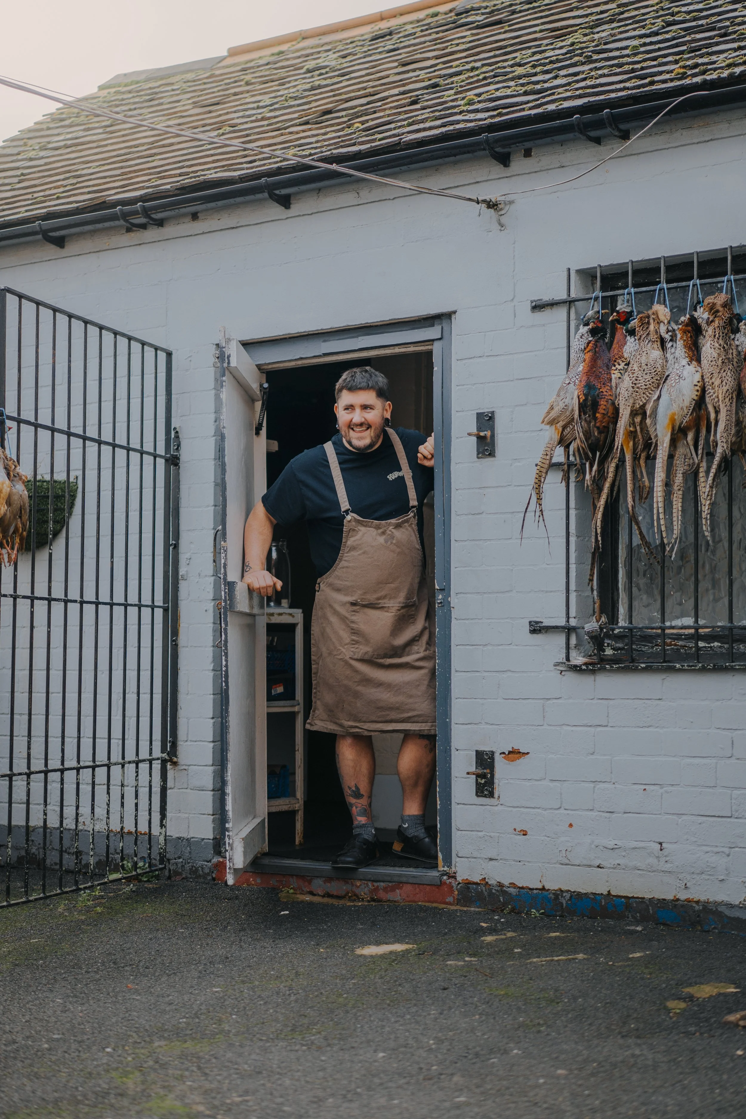 A smiling man with tattoos, wearing an apron, tweeting from a doorway outside of a white brick building with bird hides hanging on the wall.