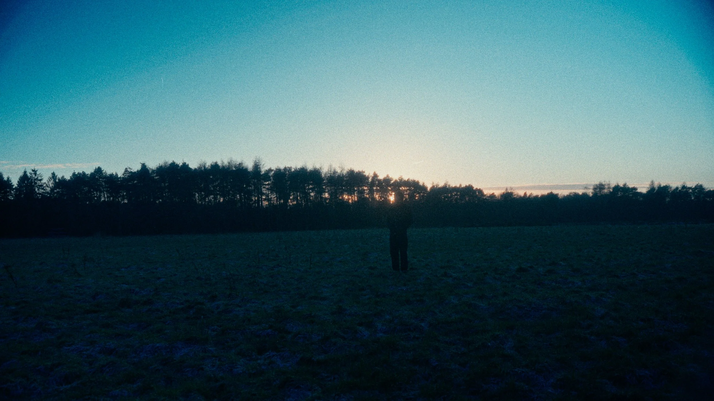 A person standing alone in a field during sunset, with a line of trees in the background and the sky transitioning from blue to orange.