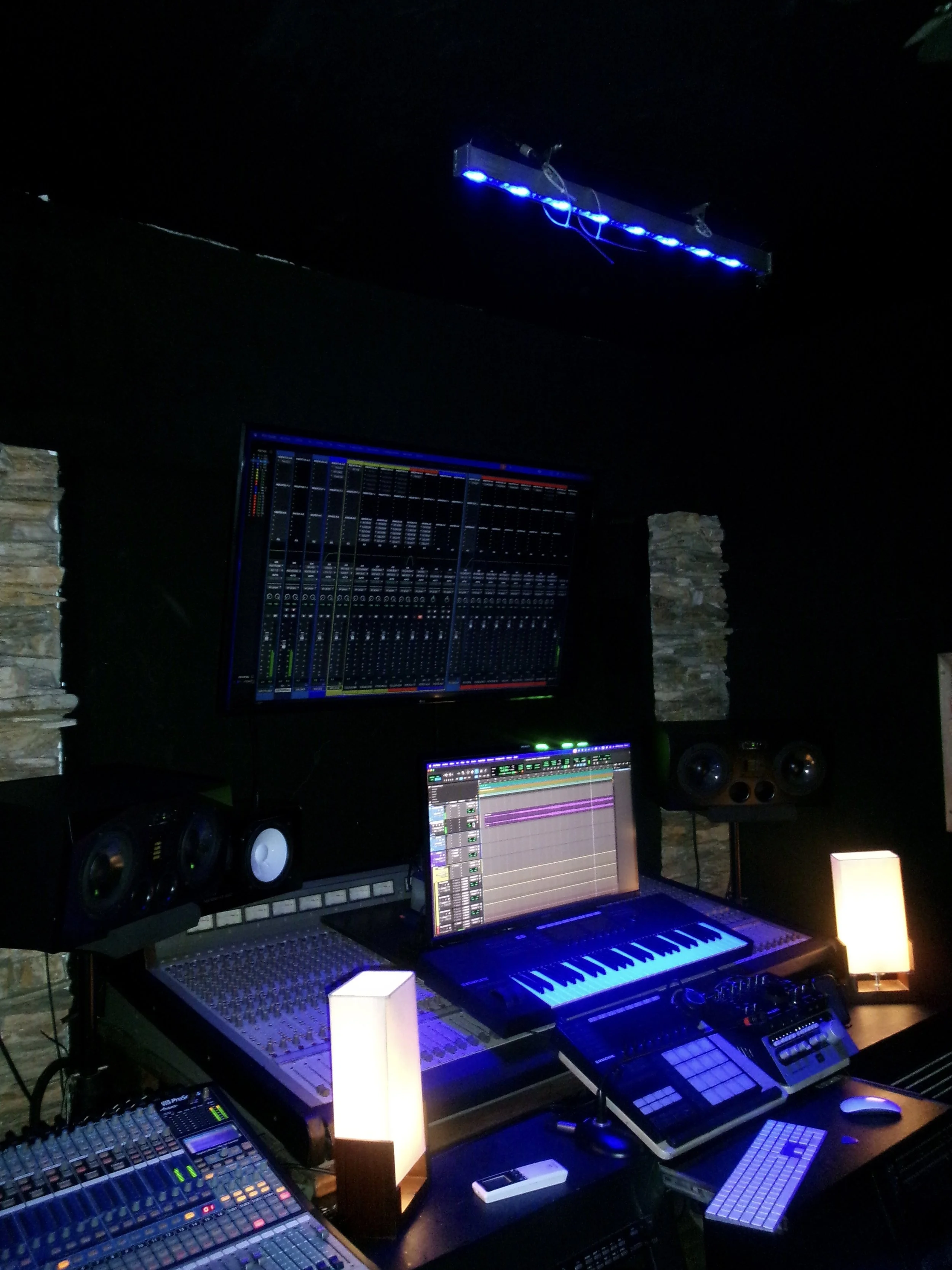 A music production studio with multiple mixing consoles, computers, and speakers illuminated by soft lighting, with stone walls and a dark ceiling.