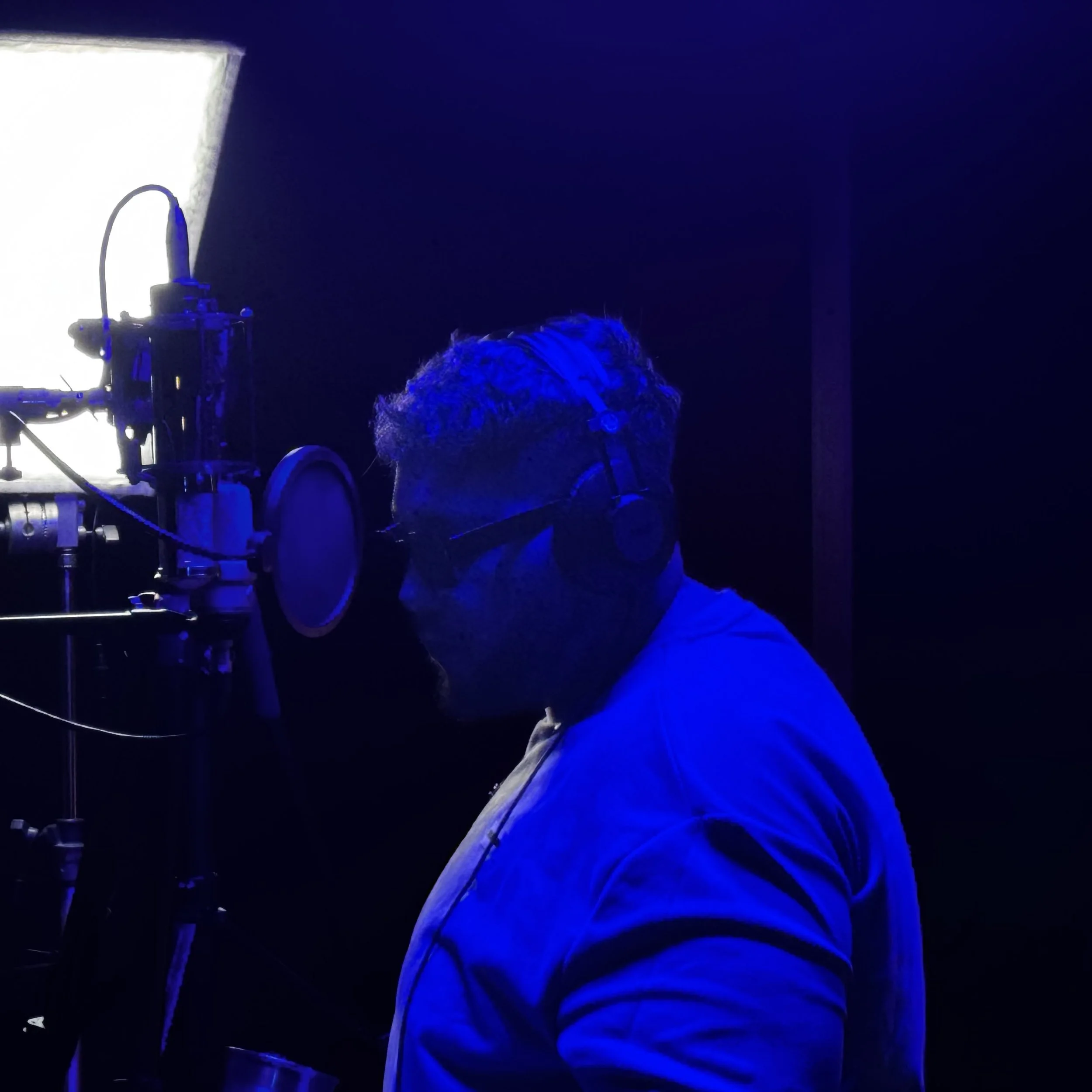 Person with curly hair wearing a white hoodie in a dark room illuminated by blue and purple light, looking at a camera setup on a stage or studio.