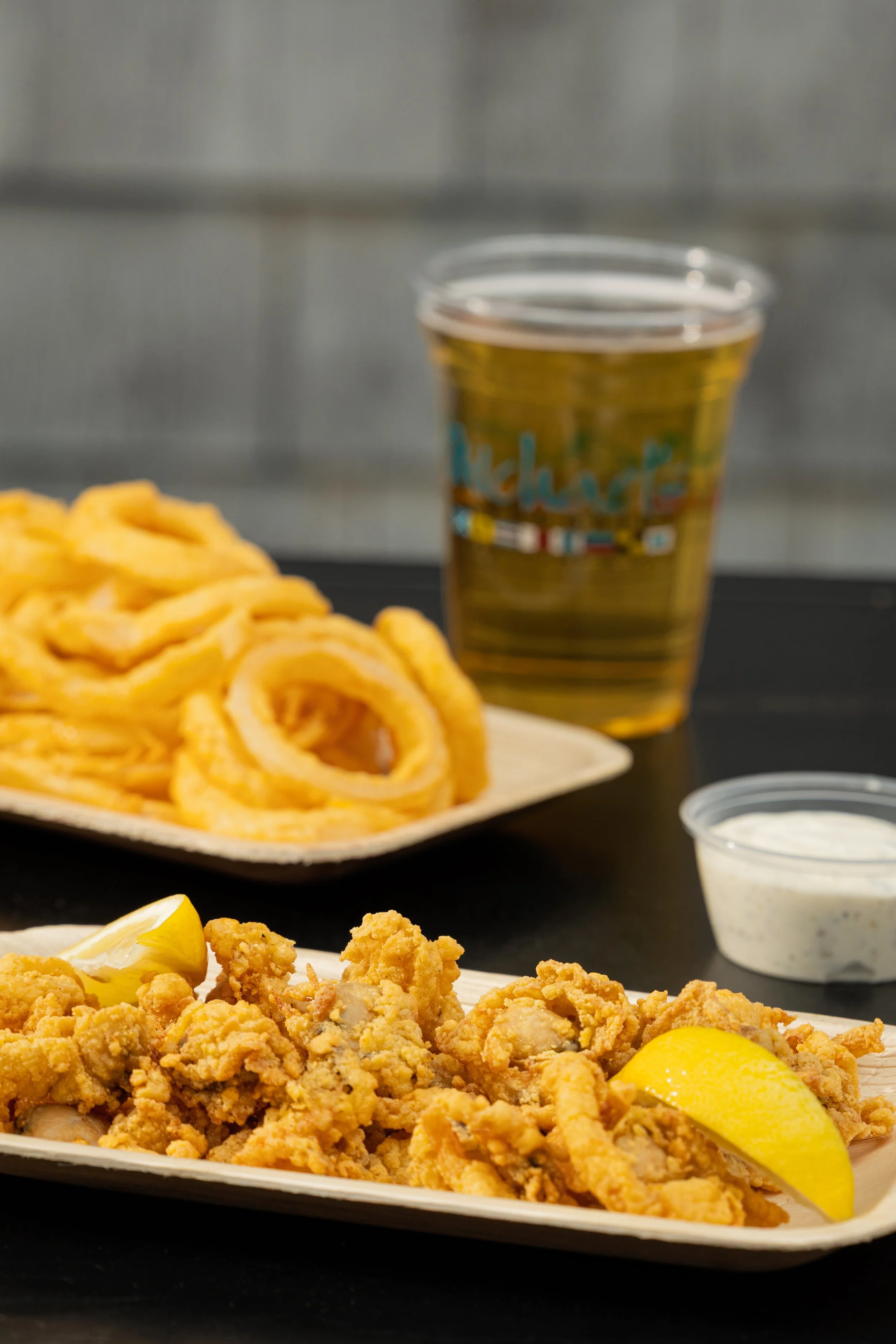 Close-up of a plate of fried food, with a lemon wedge, and a plastic cup of beer in the background, with curly fries and a side of dipping sauce.