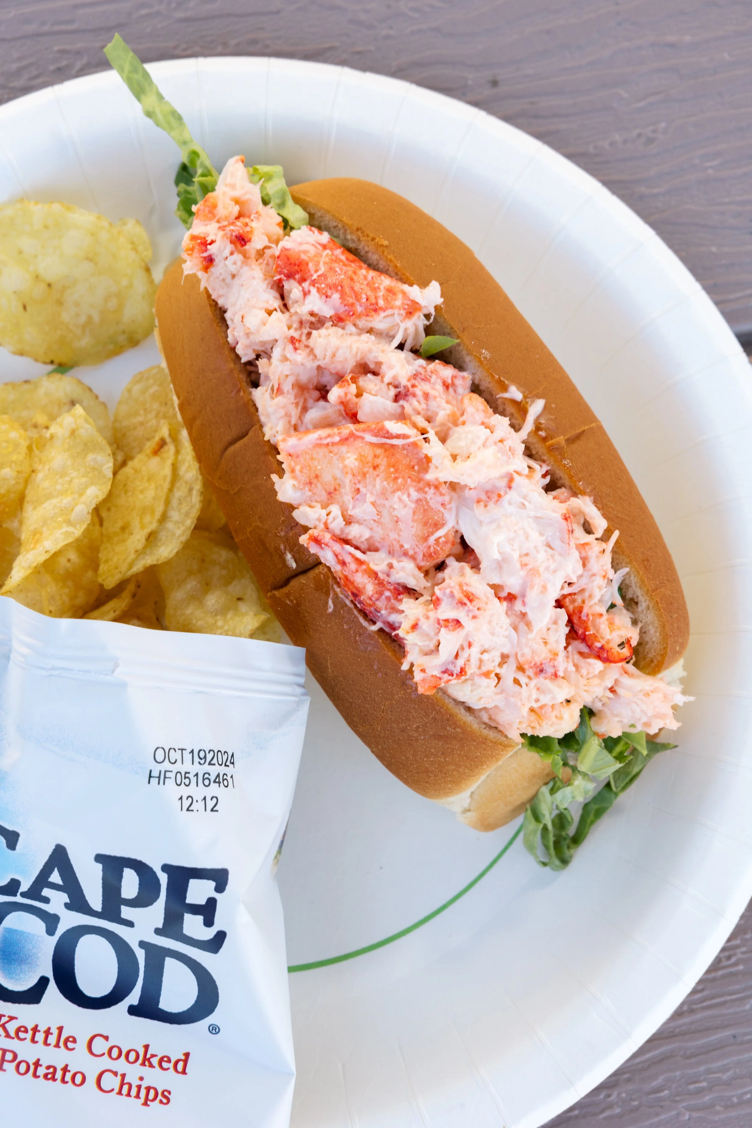 Lobster roll sandwich with potato chips on a white paper plate.