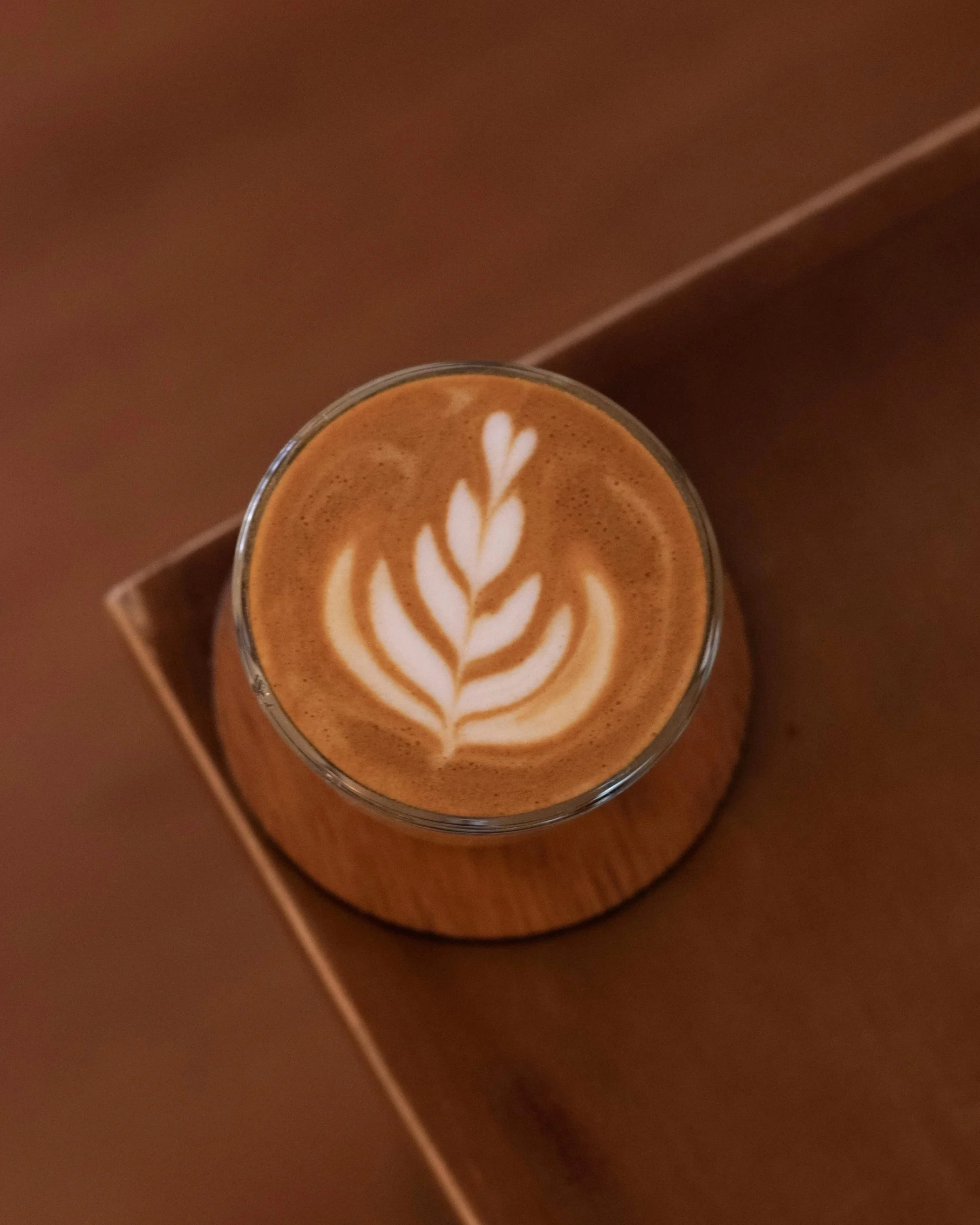 A glass of latte with a latte art leaf pattern on top, placed on a wooden coaster.