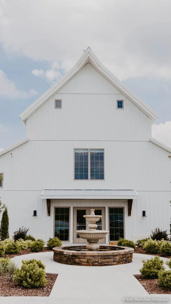 White barn with a two-tiered fountain in front, surrounded by shrubs and plants, under a partly cloudy sky.