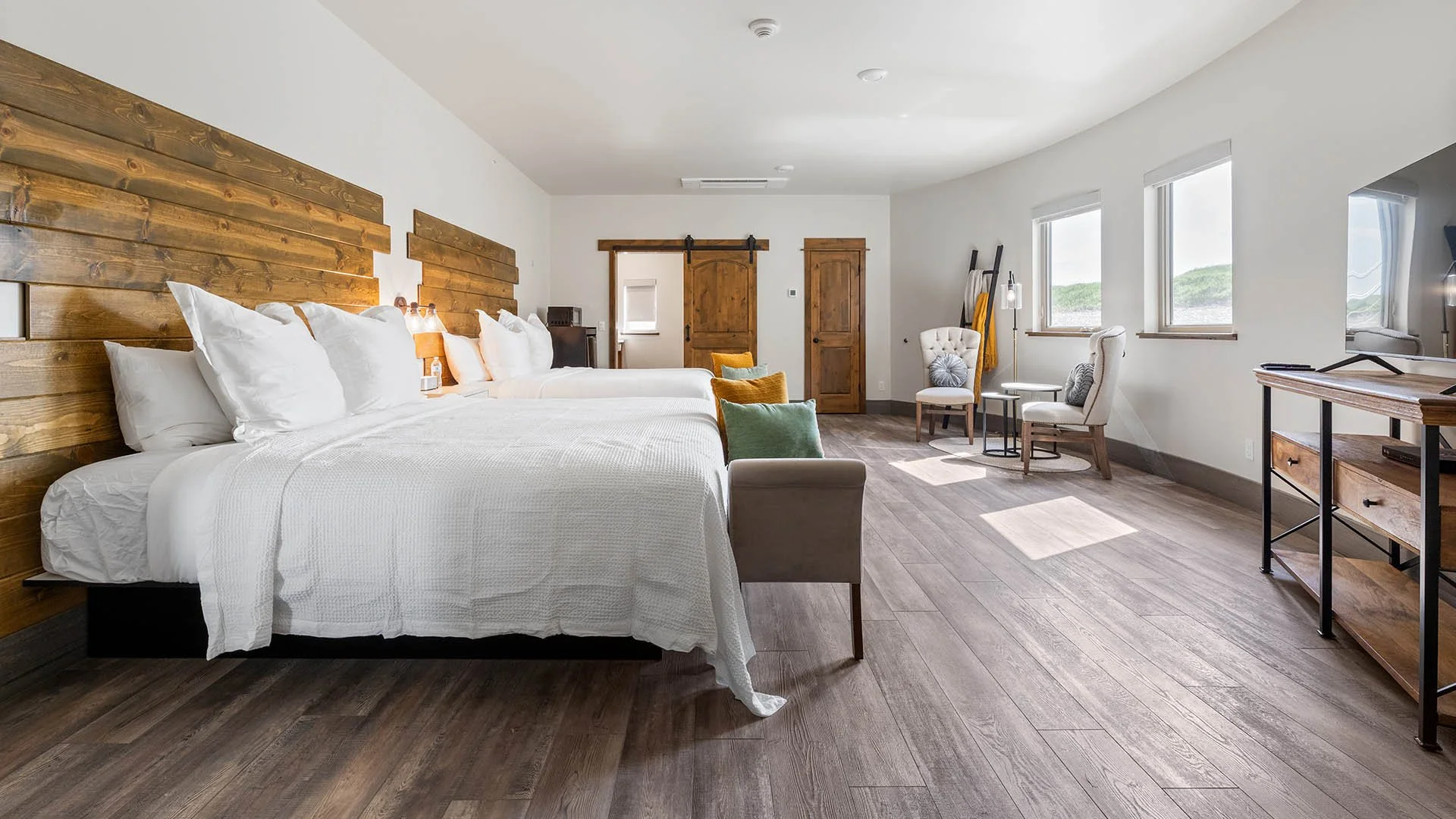 Bright hotel room with two large beds, white bedding, wooden headboards, and pillows. Features a sitting area with chairs and colorful cushions near three windows showing green hills. Wooden sliding barn doors and a TV stand with a flat-screen TV are also visible.