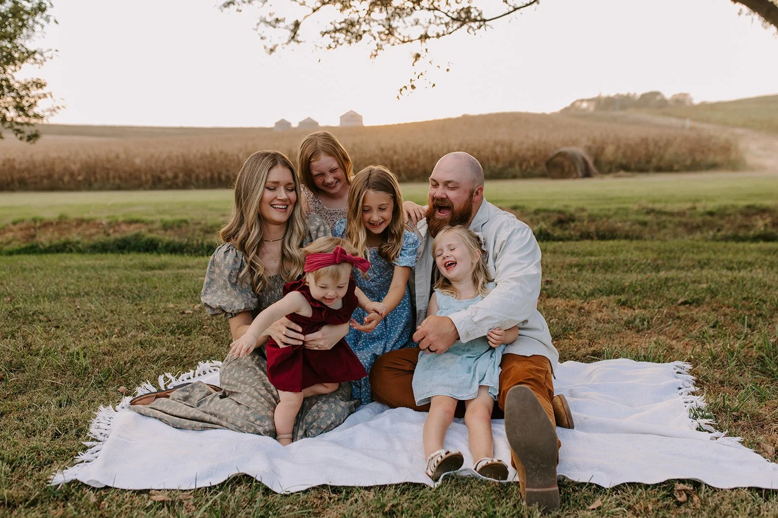 Family of seven, including children and adults, sitting on a white blanket outdoors in a field with a rural landscape, trees, and hills in the background, enjoying a sunny day.