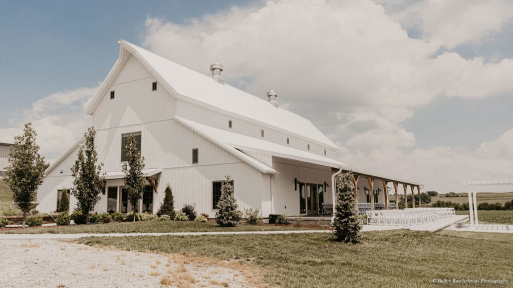 A large white barn with a gabled roof and small windows, surrounded by trees and a green lawn, under a partly cloudy sky.