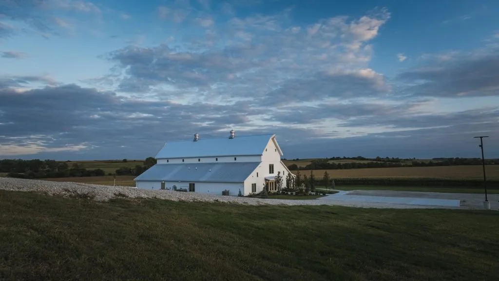 A white barn on a vast farm field with a parking area nearby, under a cloudy sky during daytime.