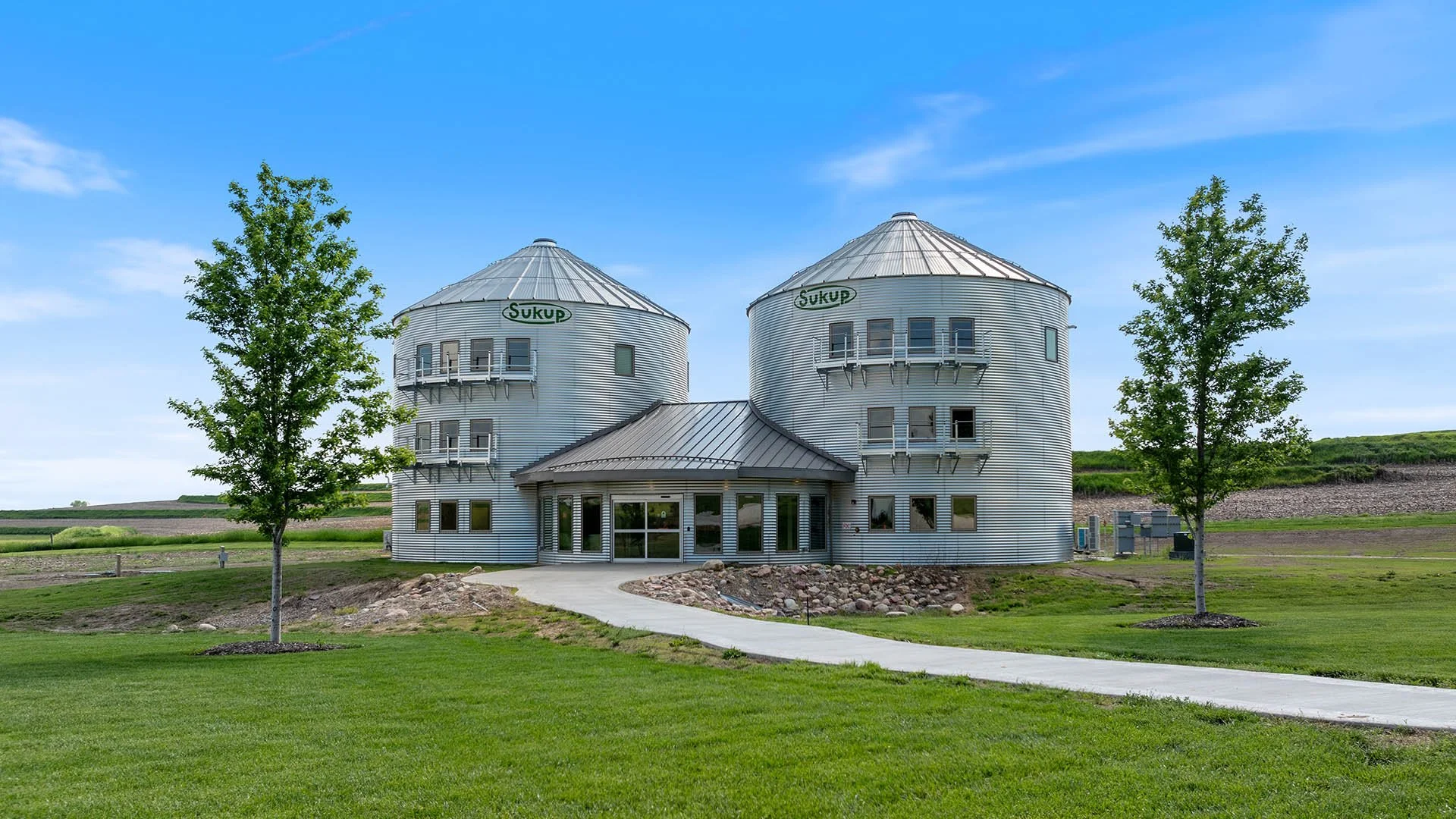 A building with two large round silos in a rural area, labeled 'SUKUP', with a curved walkway leading to the entrance, surrounded by green grass and trees under a blue sky.
