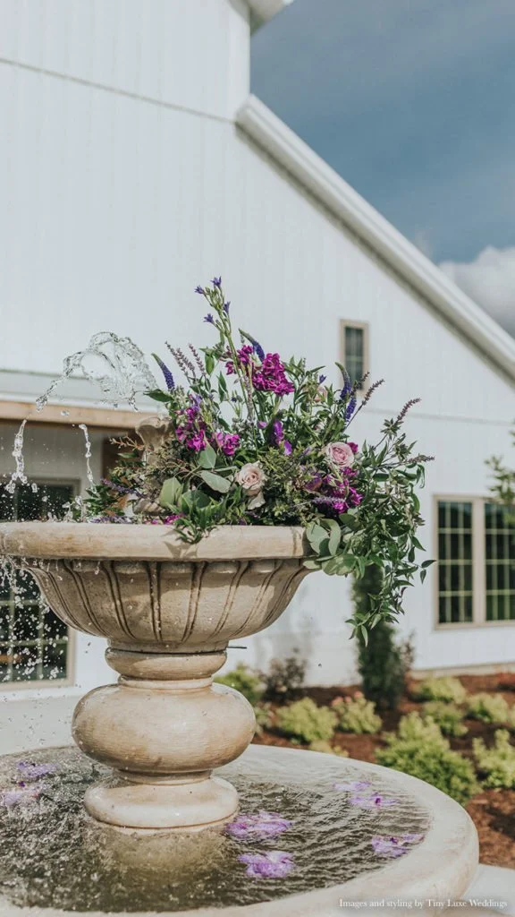 A stone water fountain decorated with a bouquet of purple, pink, and white flowers, with water flowing from the top, outside a white building with multiple windows.