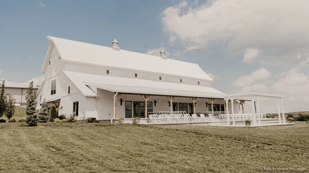 Large white barn-like building with a wraparound porch and outdoor seating, set on a grassy lawn under a partly cloudy sky.