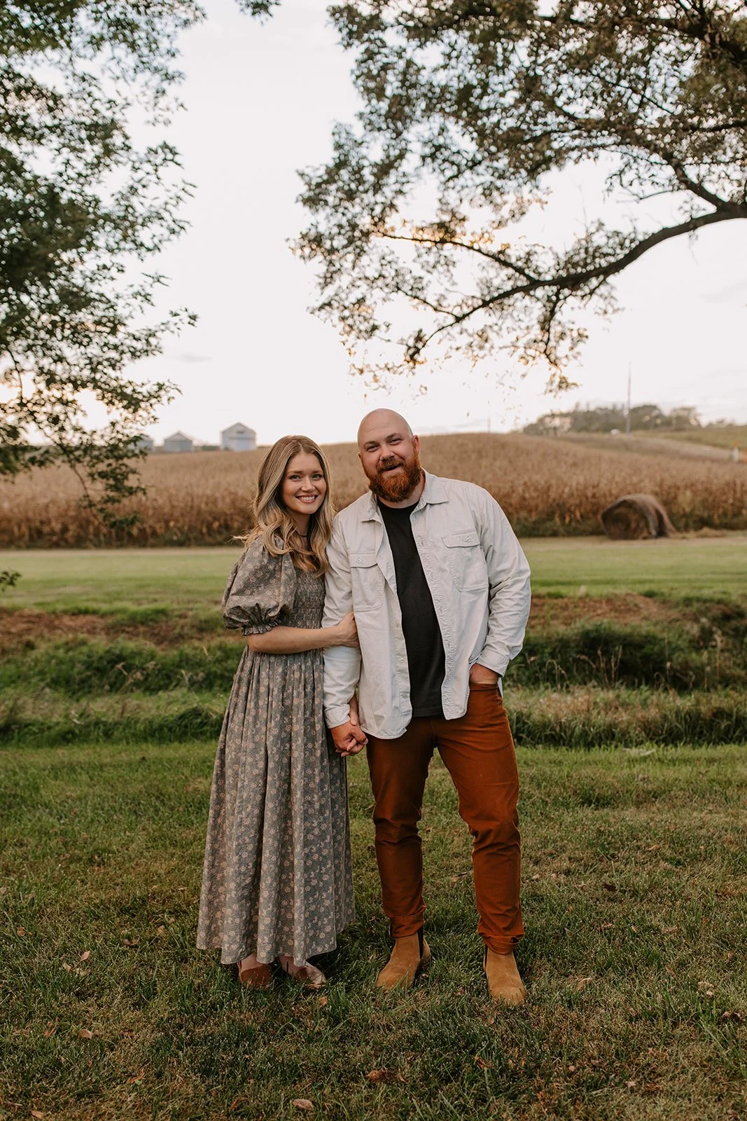 A happy couple standing hand in hand outdoors during sunset, with a field and farm buildings in the background.