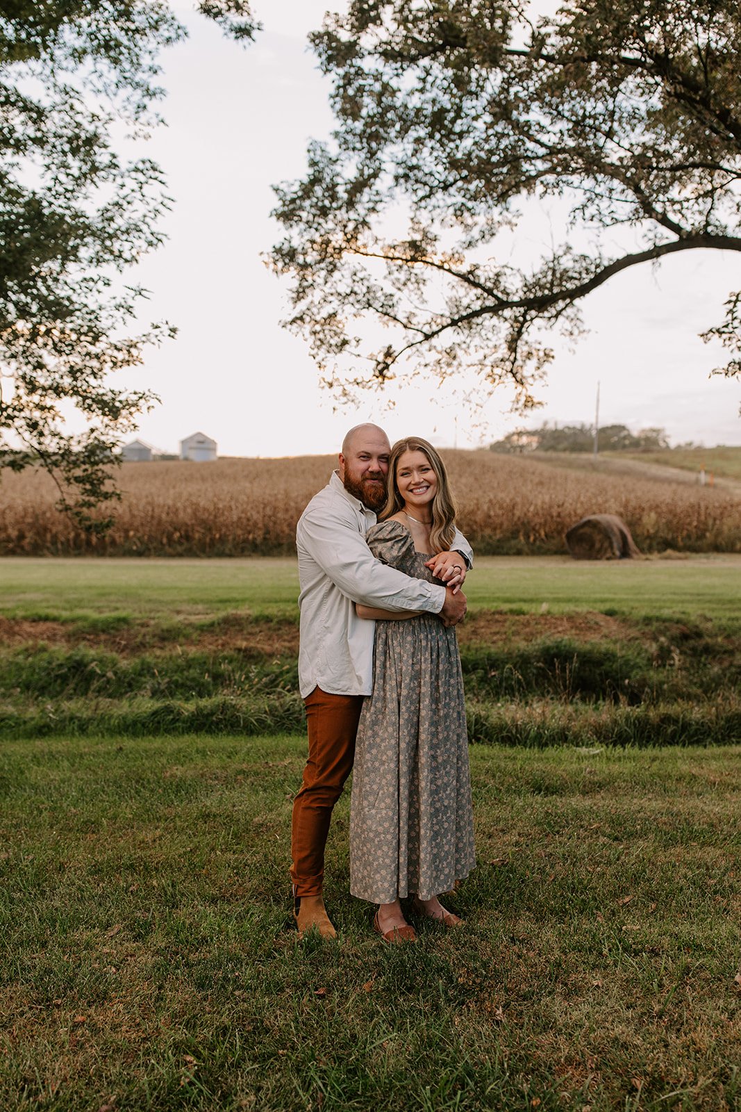 A happy couple standing outdoors on a grassy area with a farm field and large hay bale in the background during sunset, surrounded by trees.