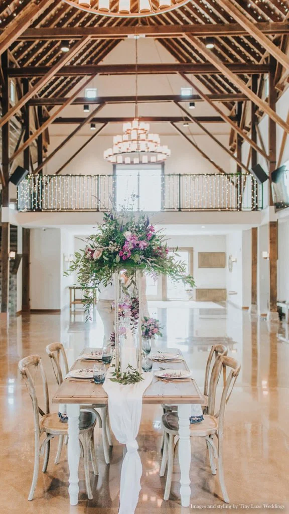 A large dining table set for a celebration in a spacious, brightly-lit barn with high wooden beams. The table has a tall floral centerpiece with purple and pink flowers, greenery, and a white cloth runner. Surrounding the table are wooden chairs, and the barn features a balcony with string lights and a chandelier hanging from the ceiling.