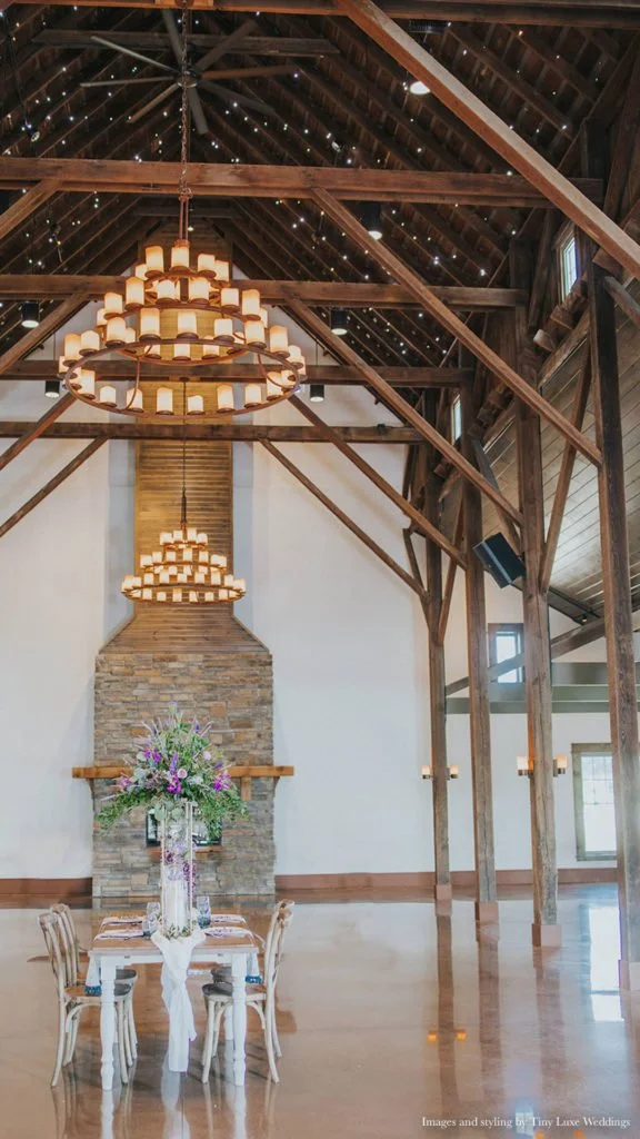 Wedding reception area with a long table, floral centerpiece, stone fireplace, wooden beams and chandeliers.