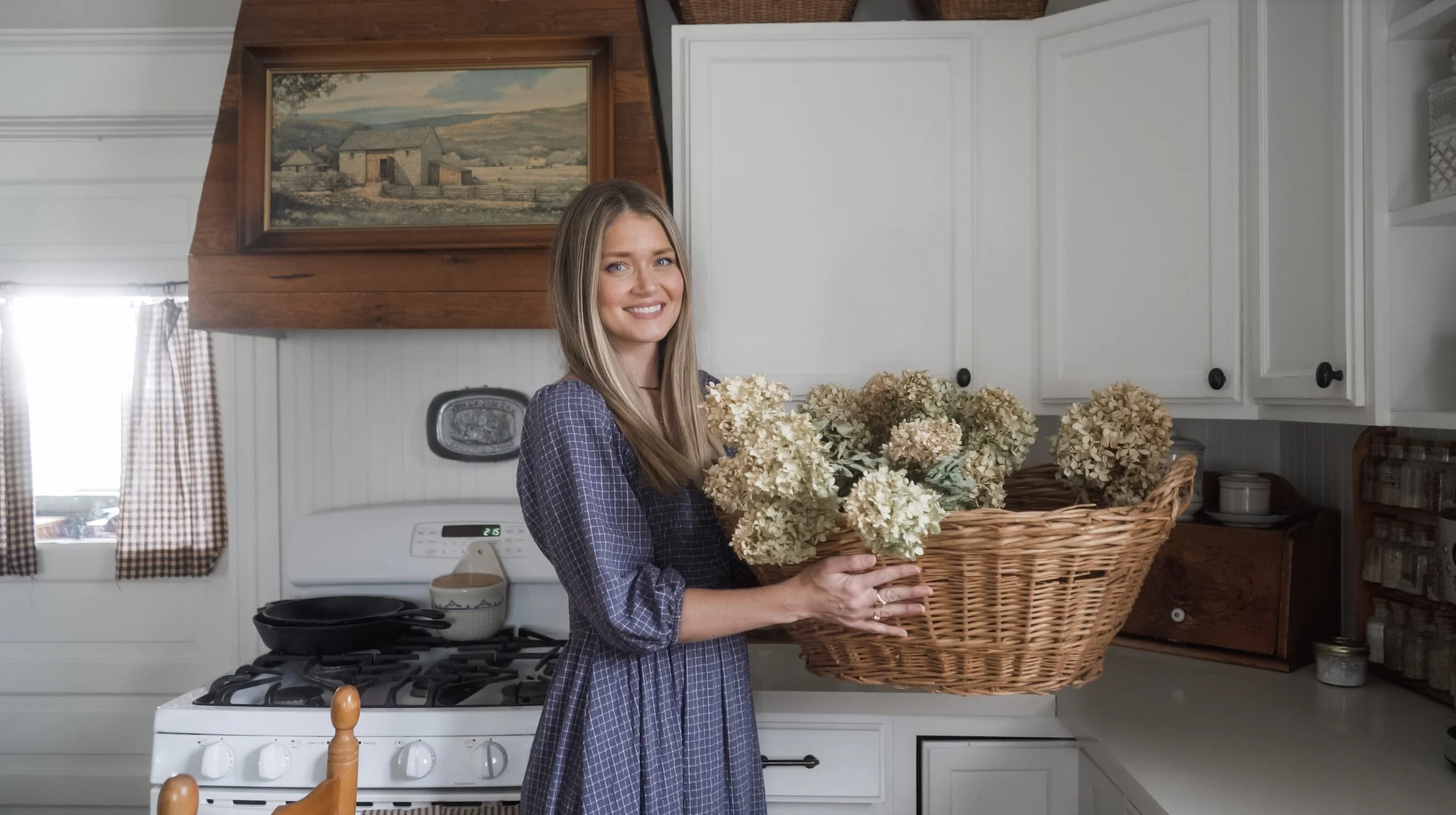 A woman with shoulder-length blonde hair wearing a blue checkered dress, standing in a kitchen holding a large basket of dried hydrangea flowers, smiling at the camera.