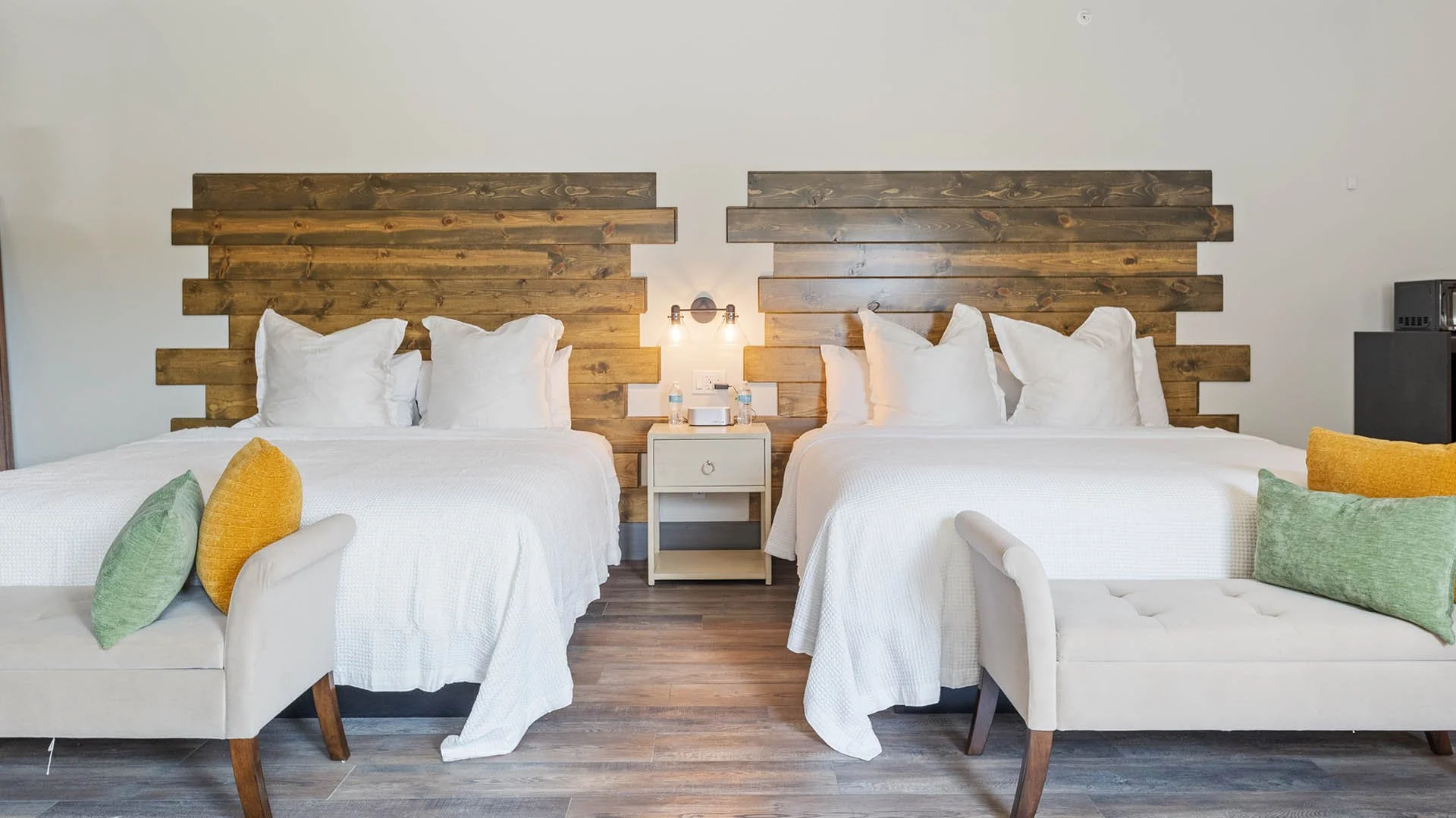 Two beds with white bedding and large pillows, with wooden headboards, a small nightstand with water bottles and an alarm clock in between, flanked by two white benches with colorful pillows, in a modern hotel room.