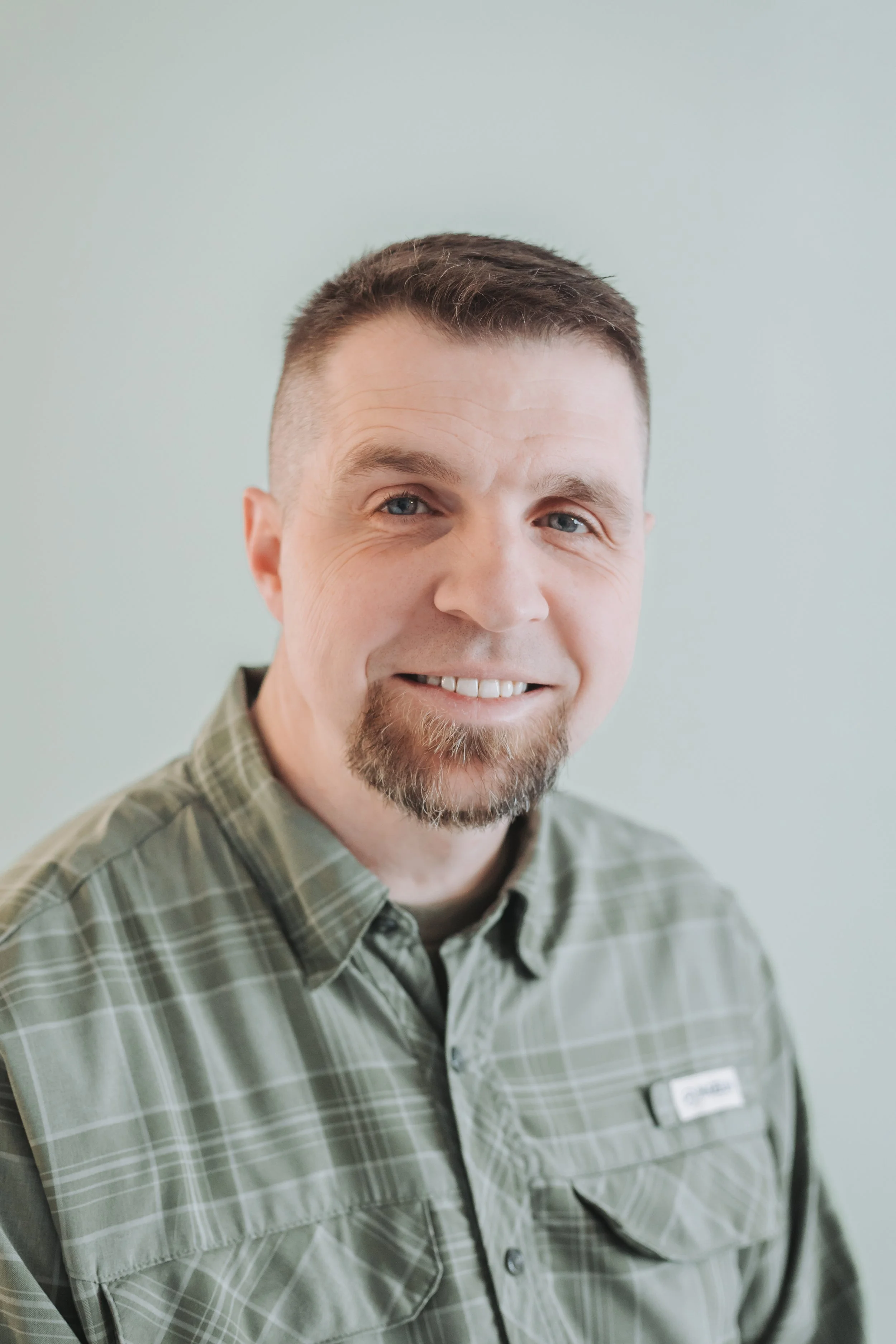 A man with short brown hair, a beard, and blue eyes smiling at the camera, wearing a green plaid shirt, against a light gray background.