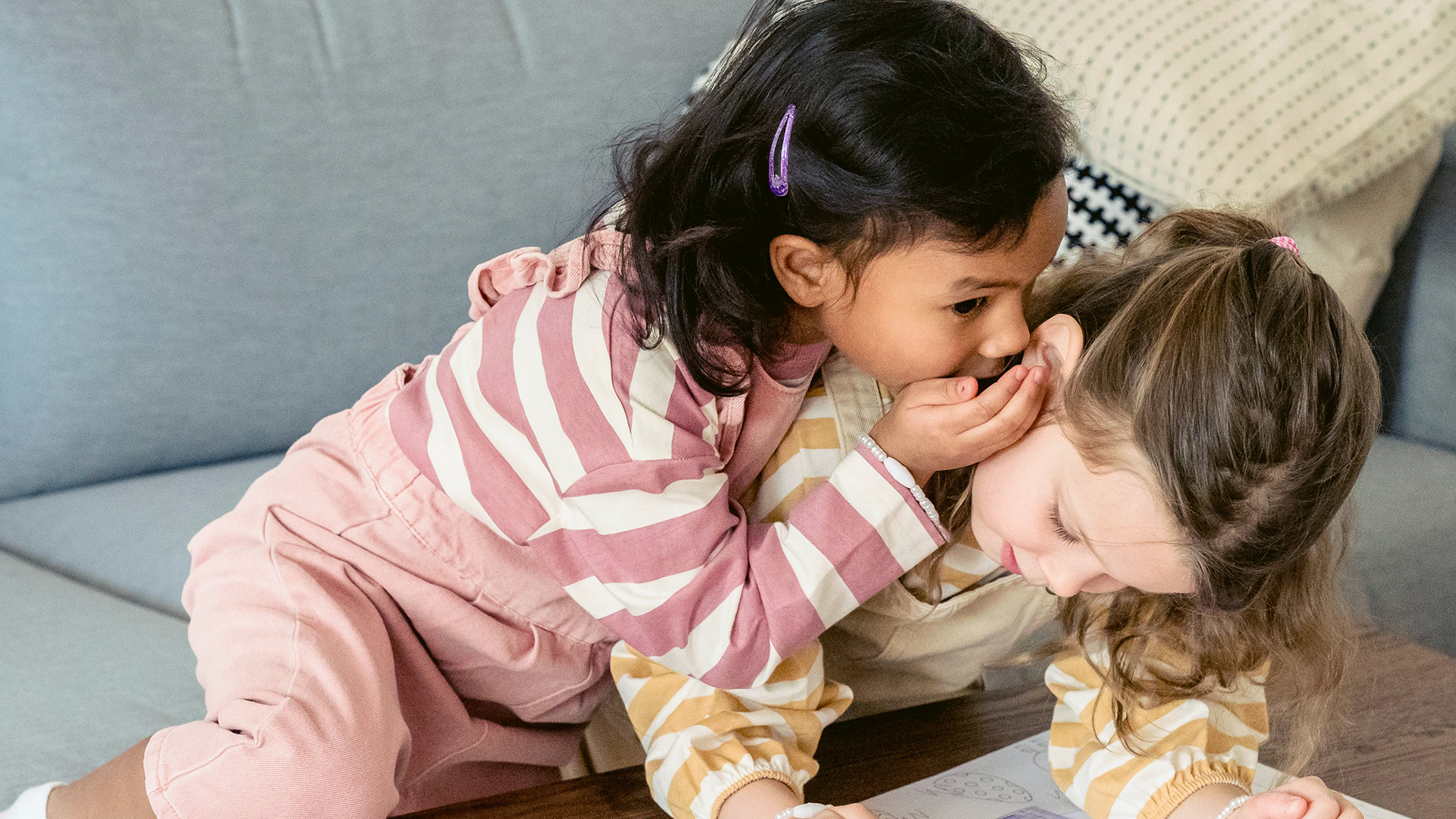 Two young toddler girls coloring and one is whispering into the other one's ear.