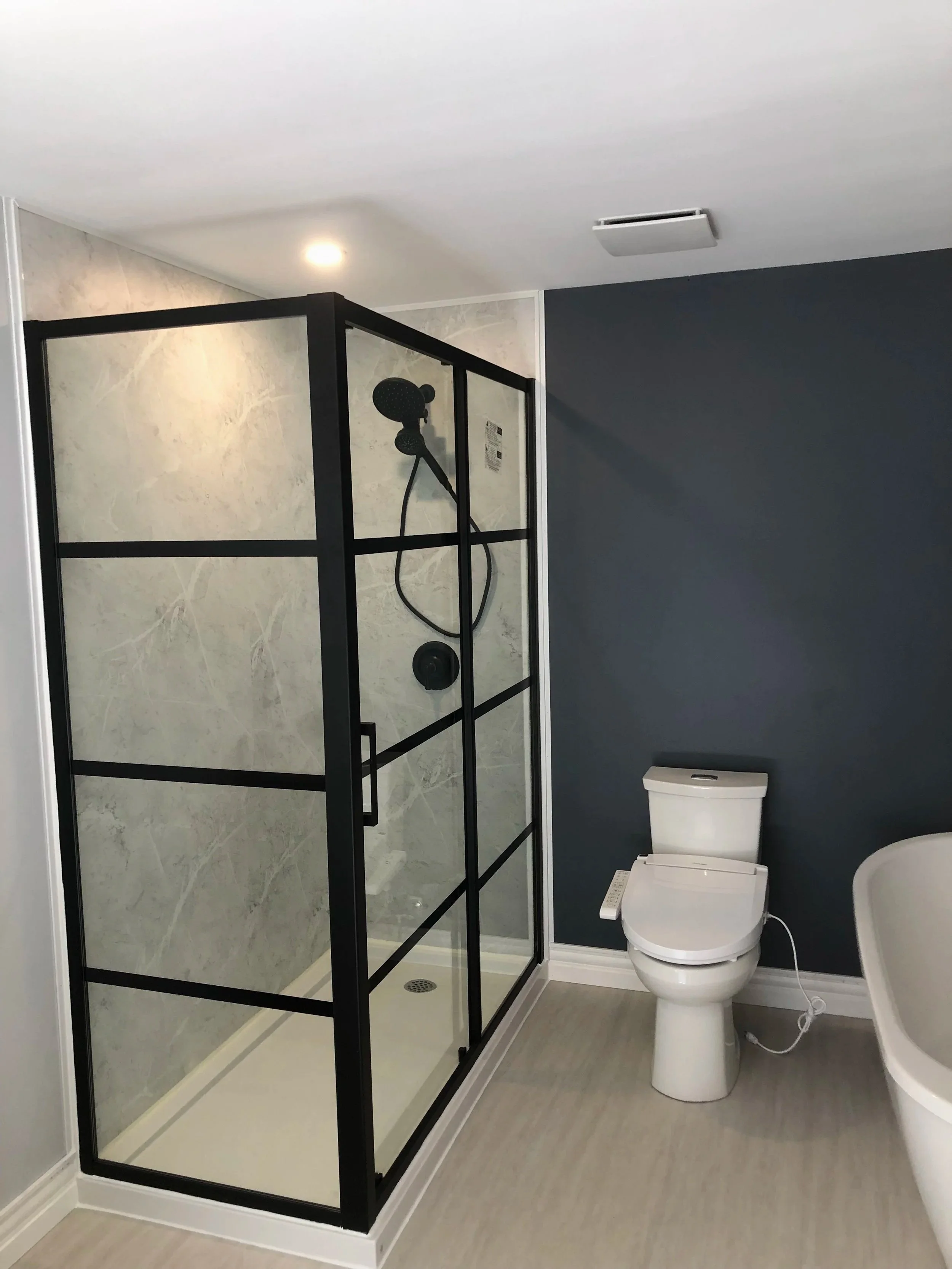 A modern bathroom with a walk-in shower with black framing, a gray interior, and a white toilet with a bidet attachment, against a dark gray wall and light-colored floor.