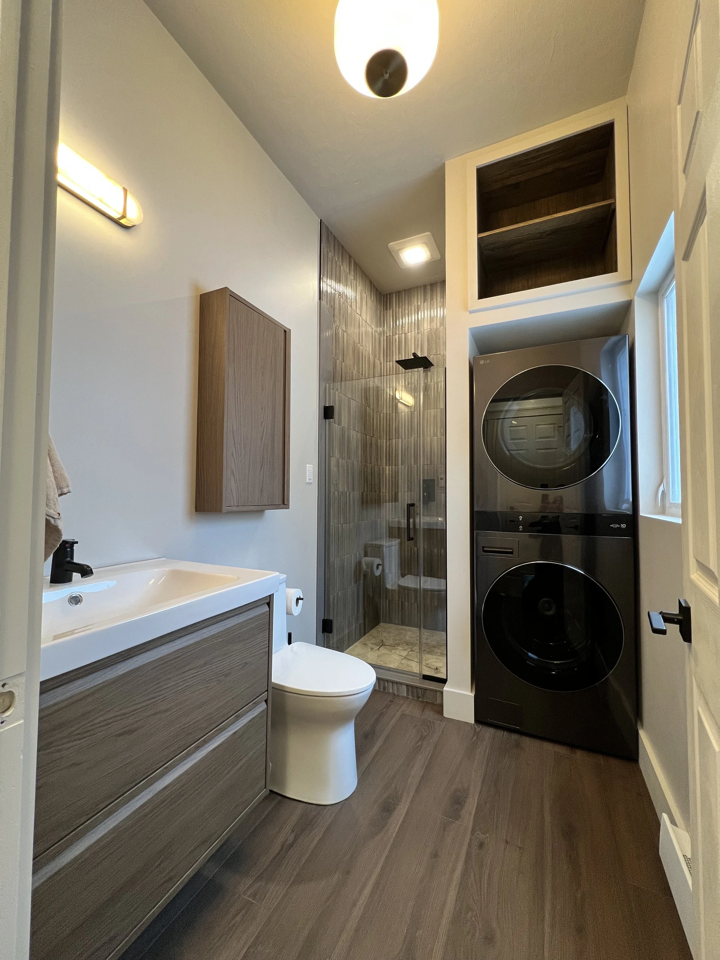 Bathroom with a shower, toilet, sink, wood flooring, stacked washer and dryer, and a small window.