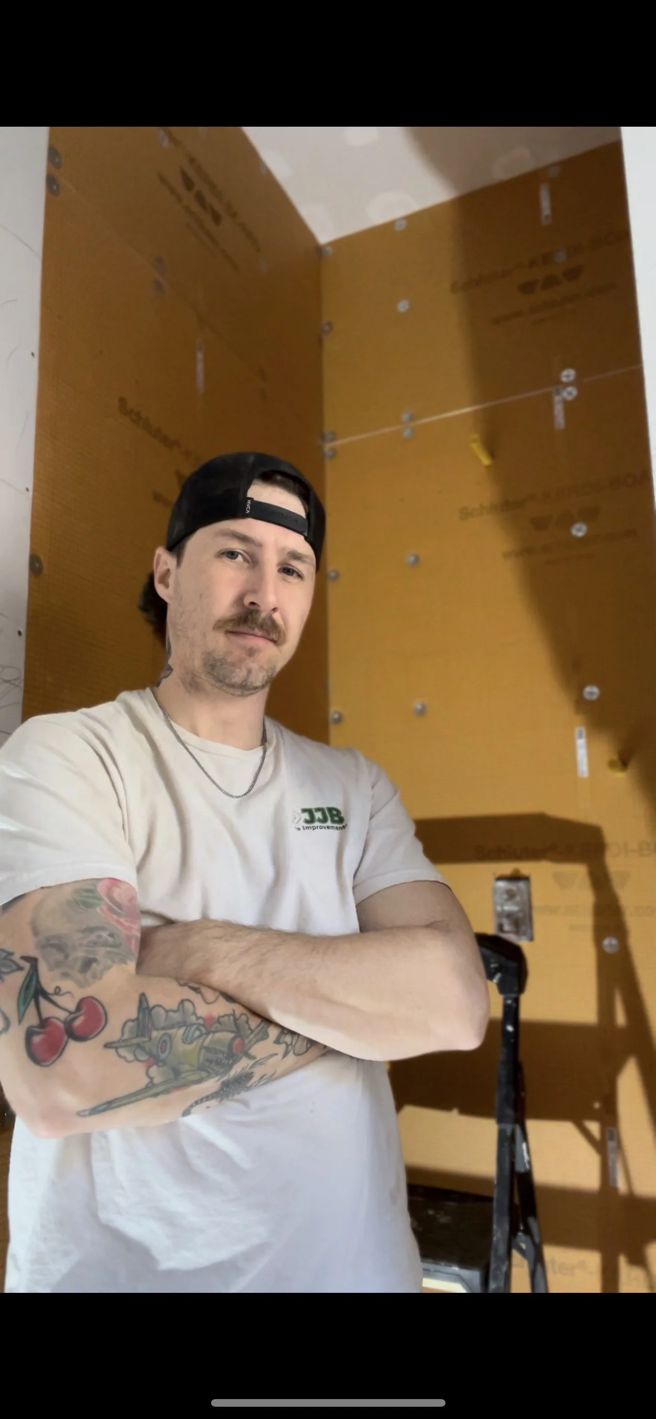 A man with tattoos and a beard wearing a white t-shirt and black backward cap, standing with arms crossed in a room with yellow drywall and metal fasteners, possibly under construction.