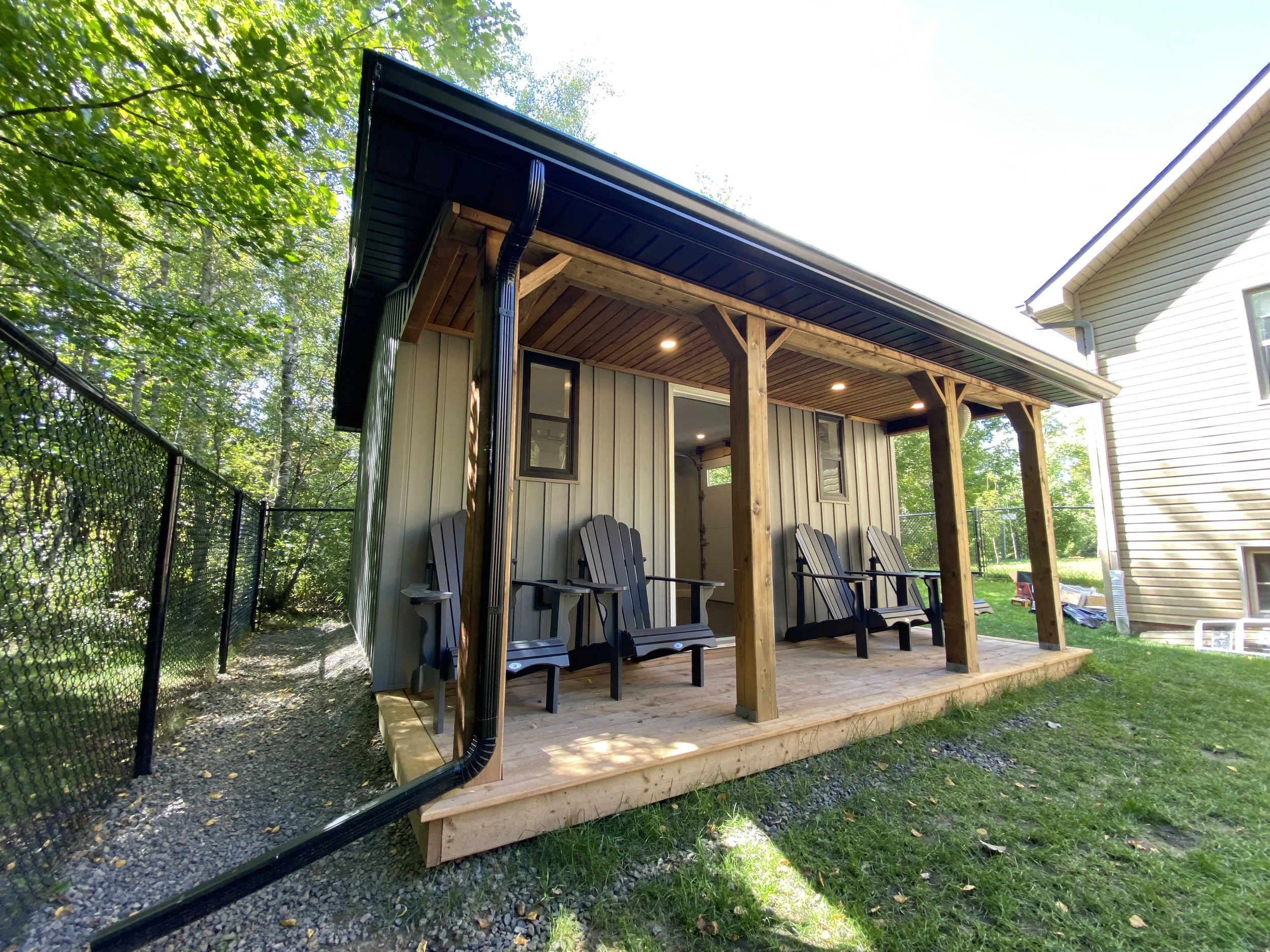 A backyard patio with four black Adirondack chairs under a wooden roof extension, attached to a beige house with trees in the background.