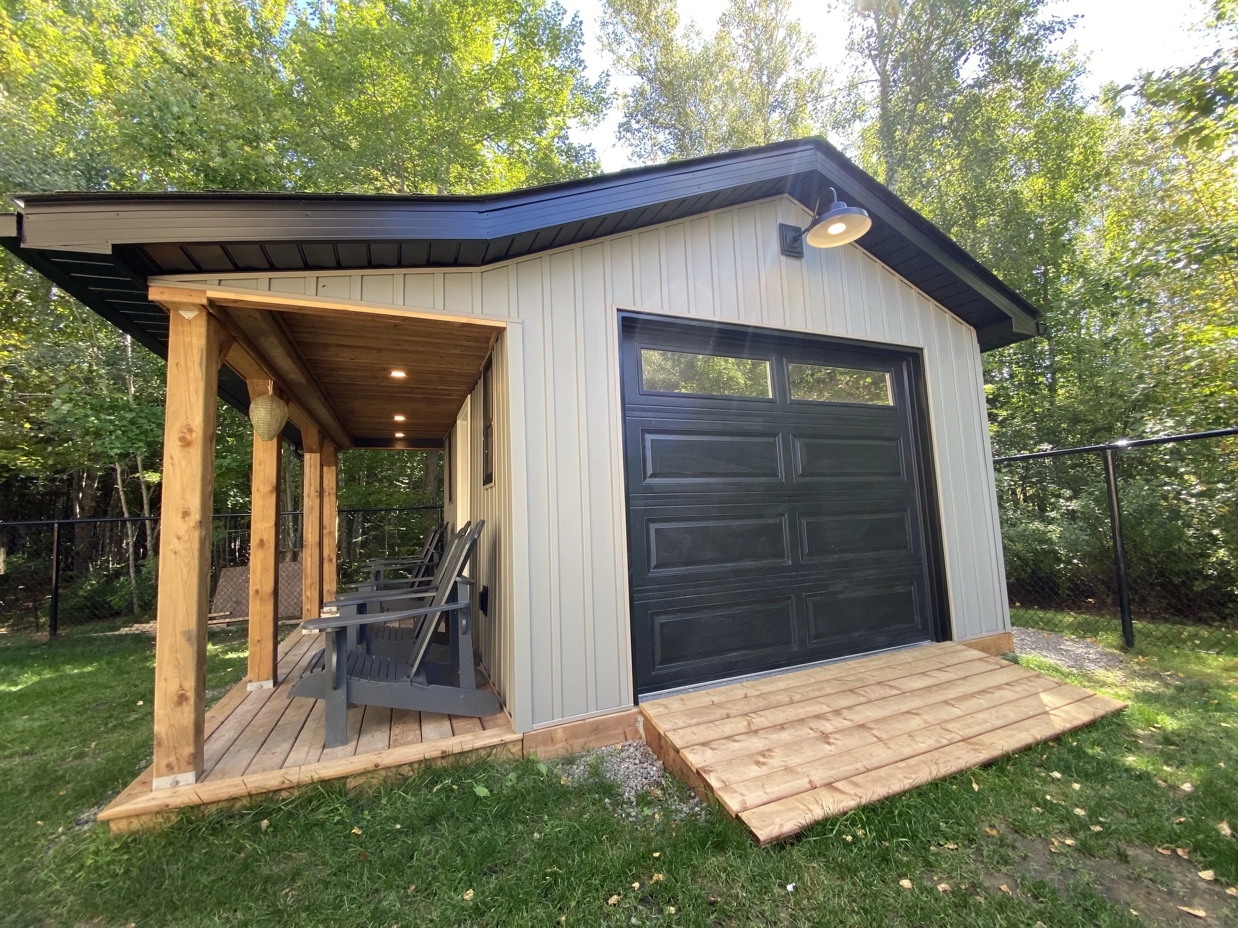 A small modern shed with a black garage door, a wood porch with outdoor seating, and surrounded by green trees.