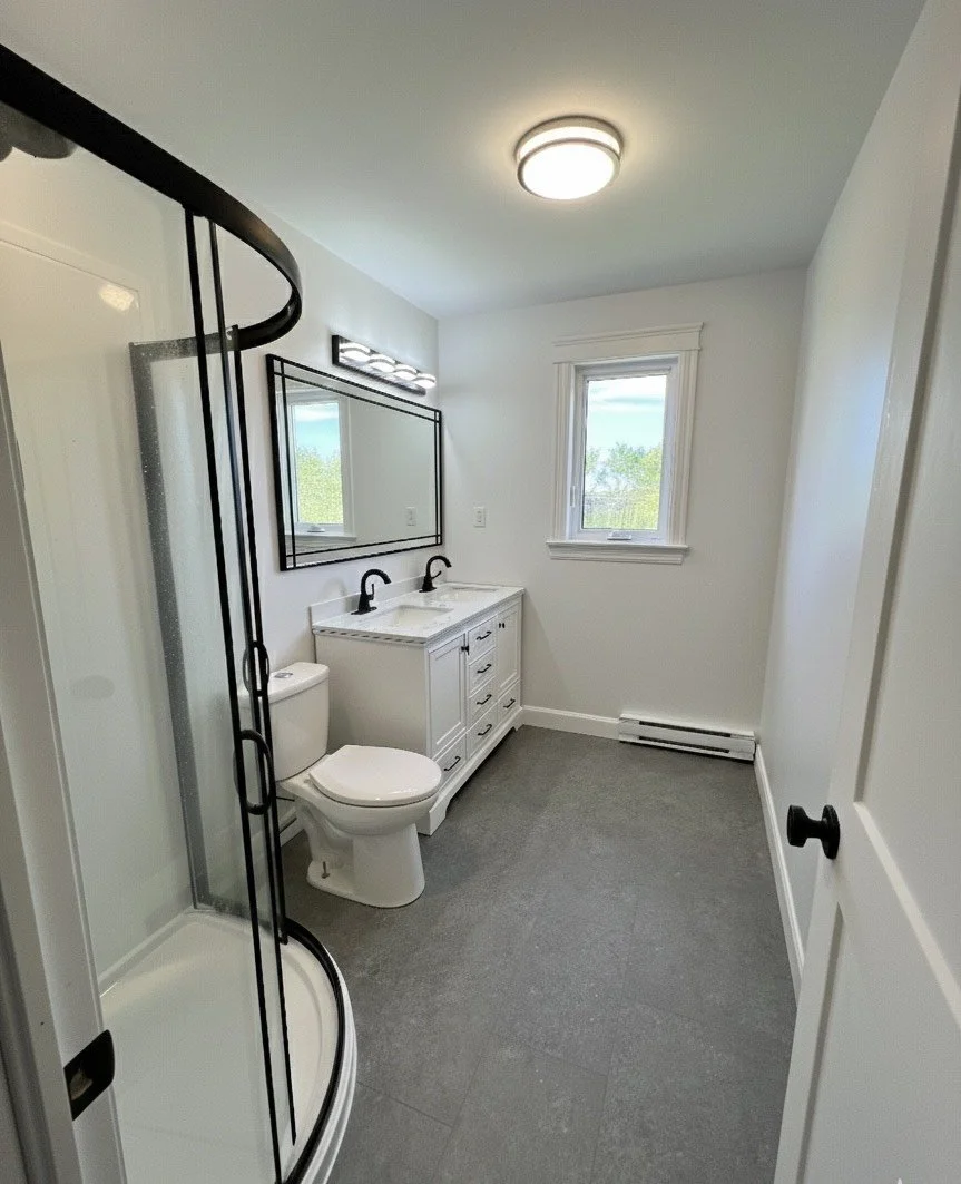 A modern bathroom with a shower stall, a white toilet, a vanity with a double sink and black faucets, a large mirror, a window showing greenery outside, a ceiling light, and a baseboard heater.