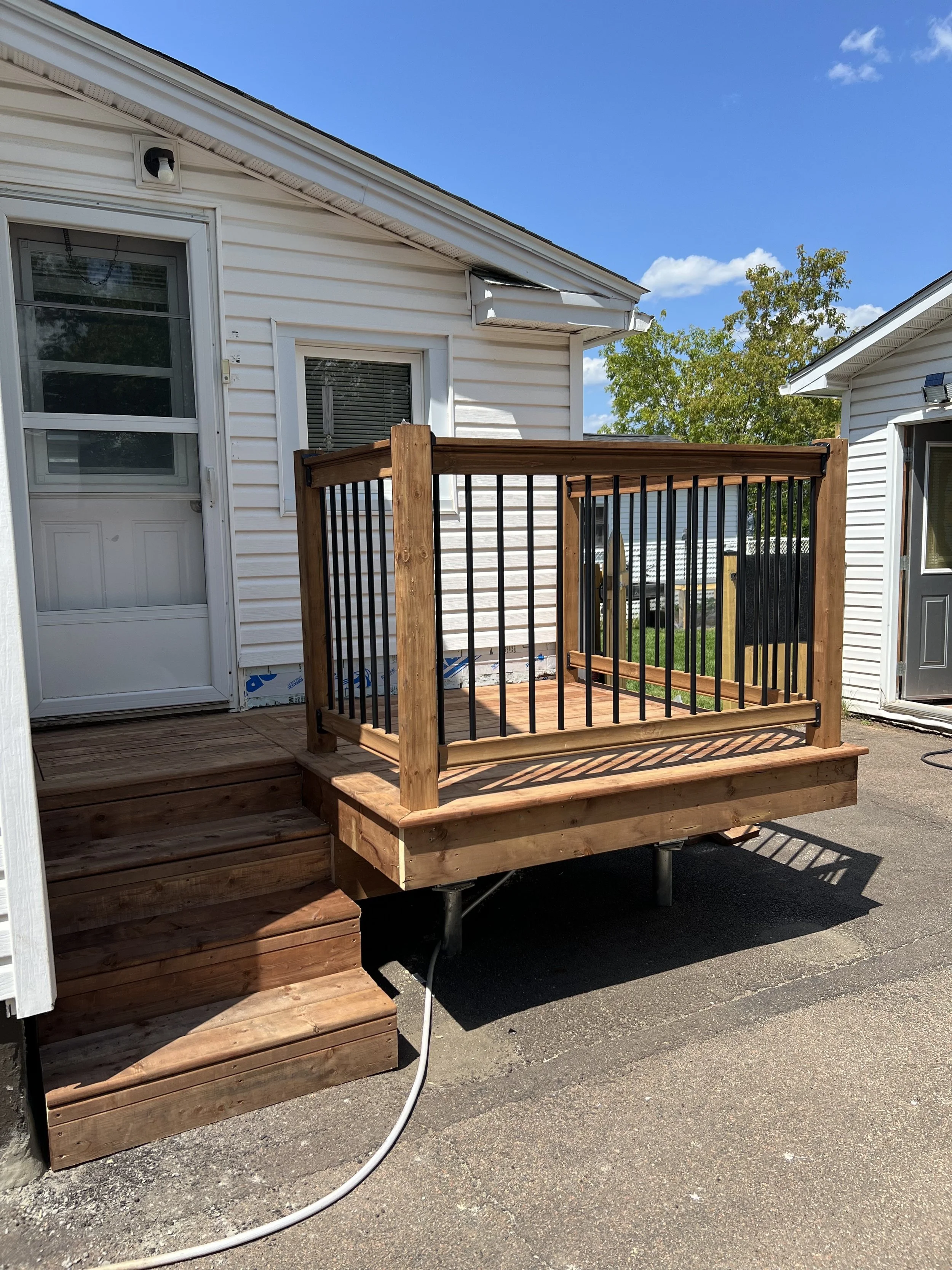 Newly built wooden deck with black metal railings outside a white house, with stairs leading up to it. The deck is situated on a paved surface on a sunny day with a blue sky and some green trees in the background.