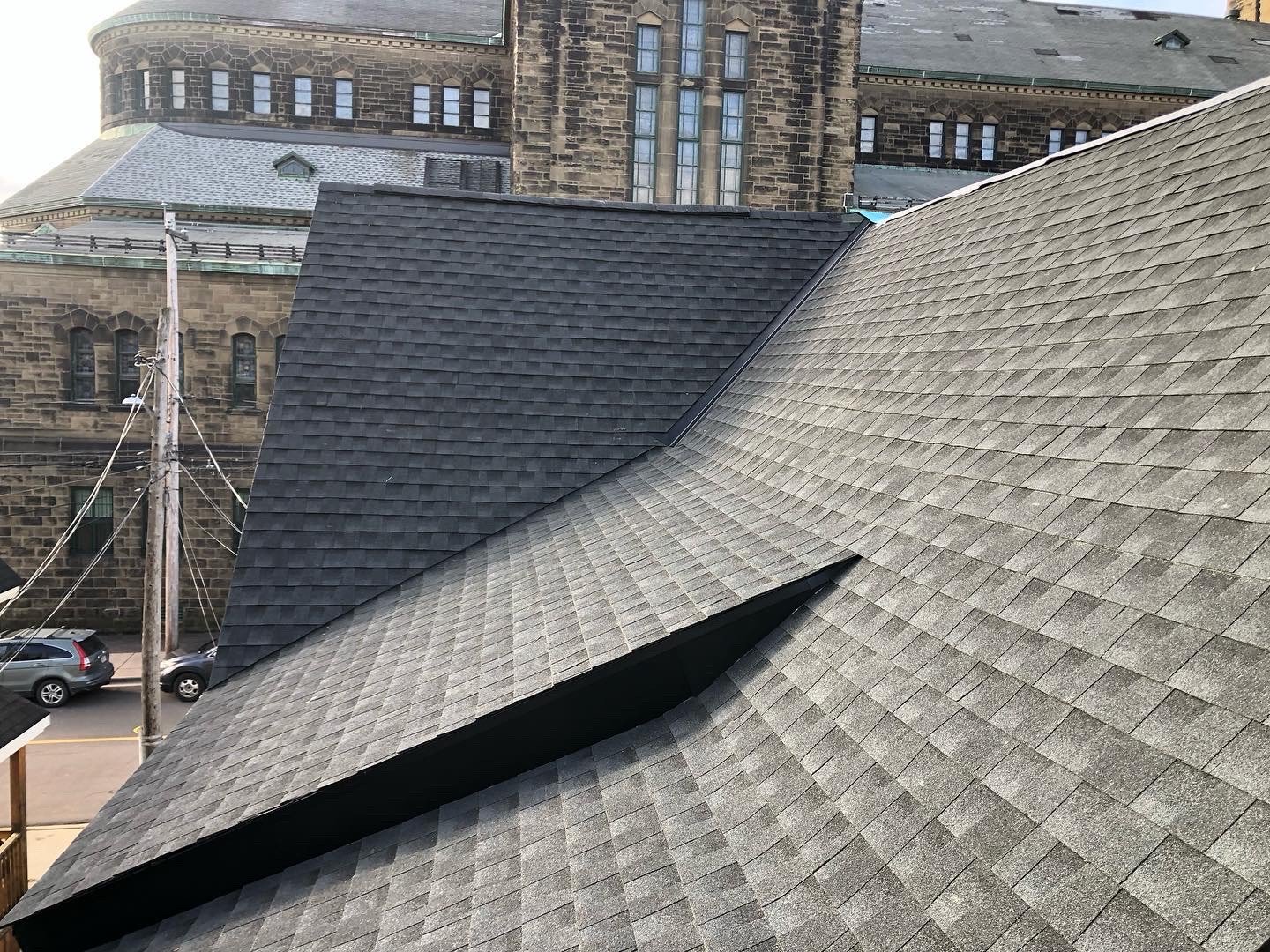 A close-up view of a rooftop with asphalt shingles in shades of gray and black, showing various slopes and angles, with a historic stone building in the background.