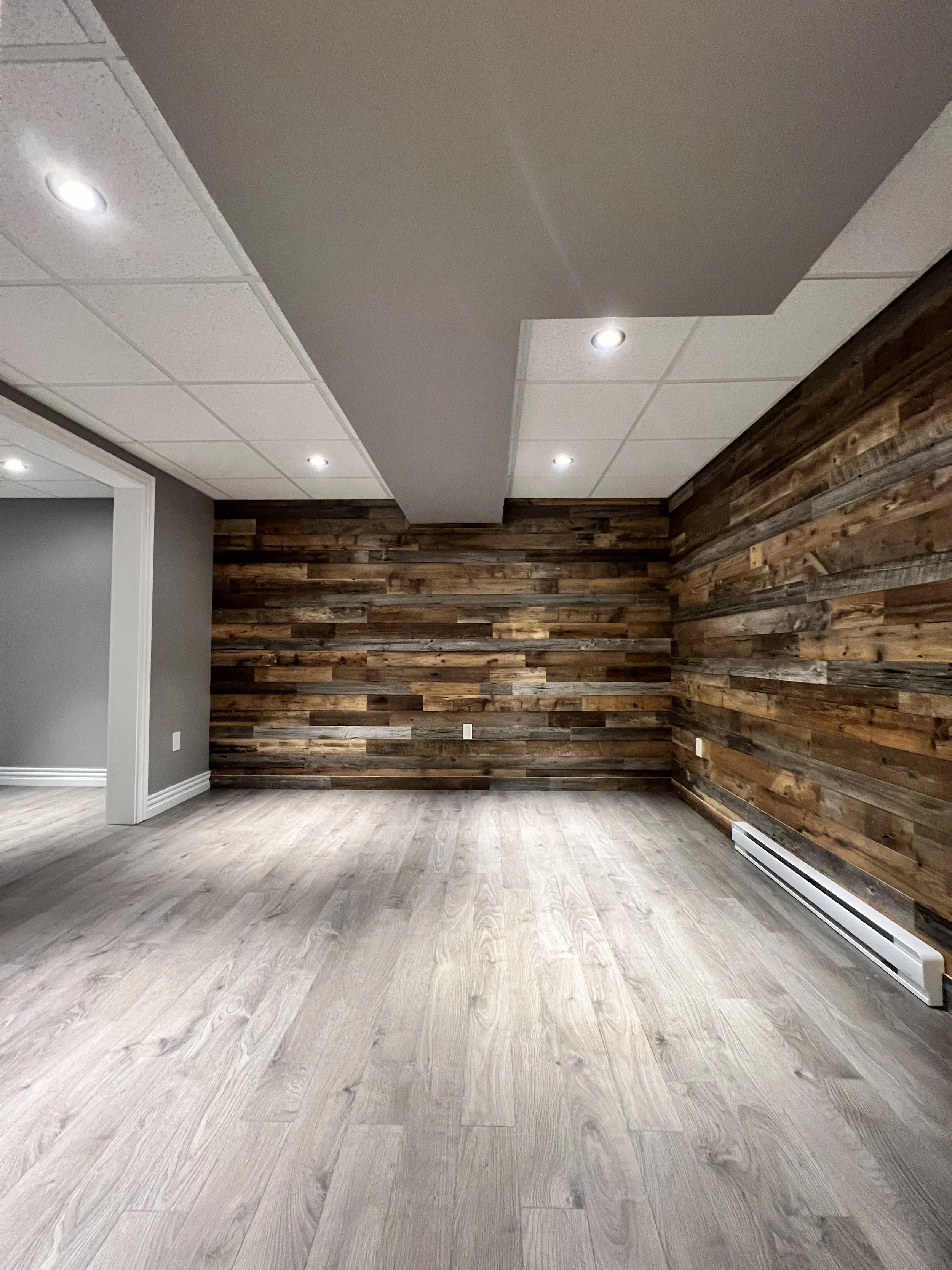 Interior room with wood-paneled wall, light-colored wood flooring, and a dropped ceiling with recessed lighting.