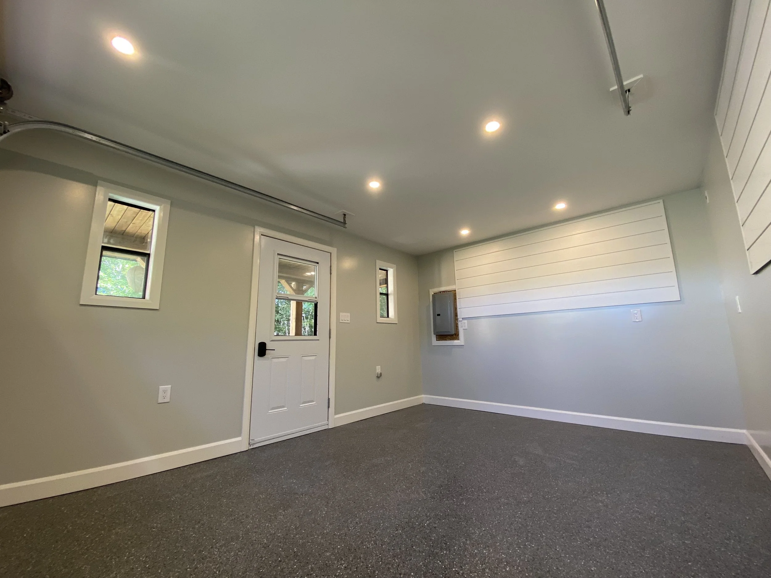 Empty room with gray carpet, white walls, and small windows, with a white door and a large white wooden pattern on one wall, and ceiling lights.
