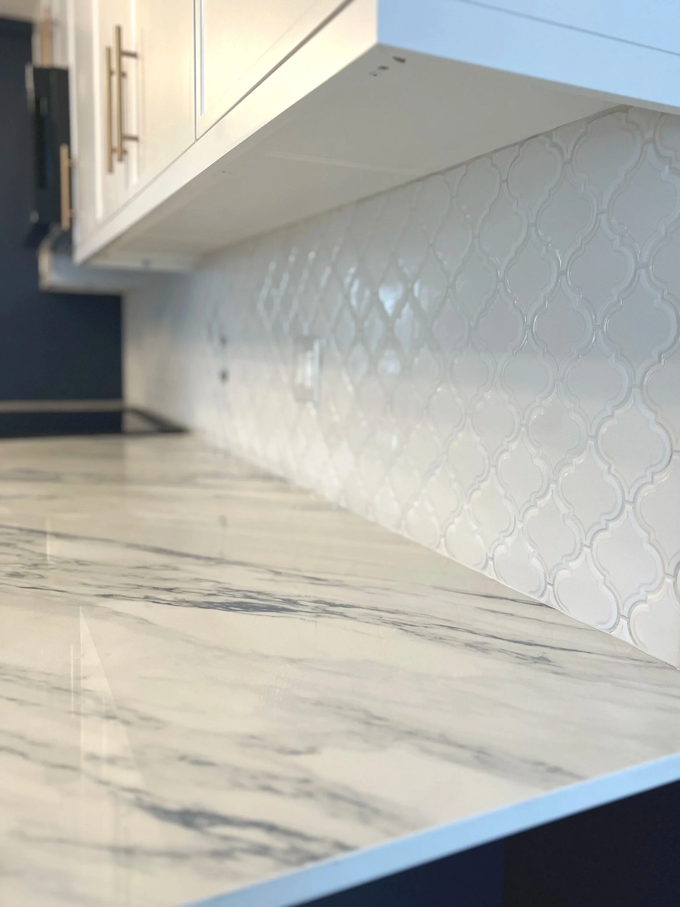 Close-up view of a white marble countertop, kitchen backsplash with white patterned tiles, white cabinetry with brass handles.