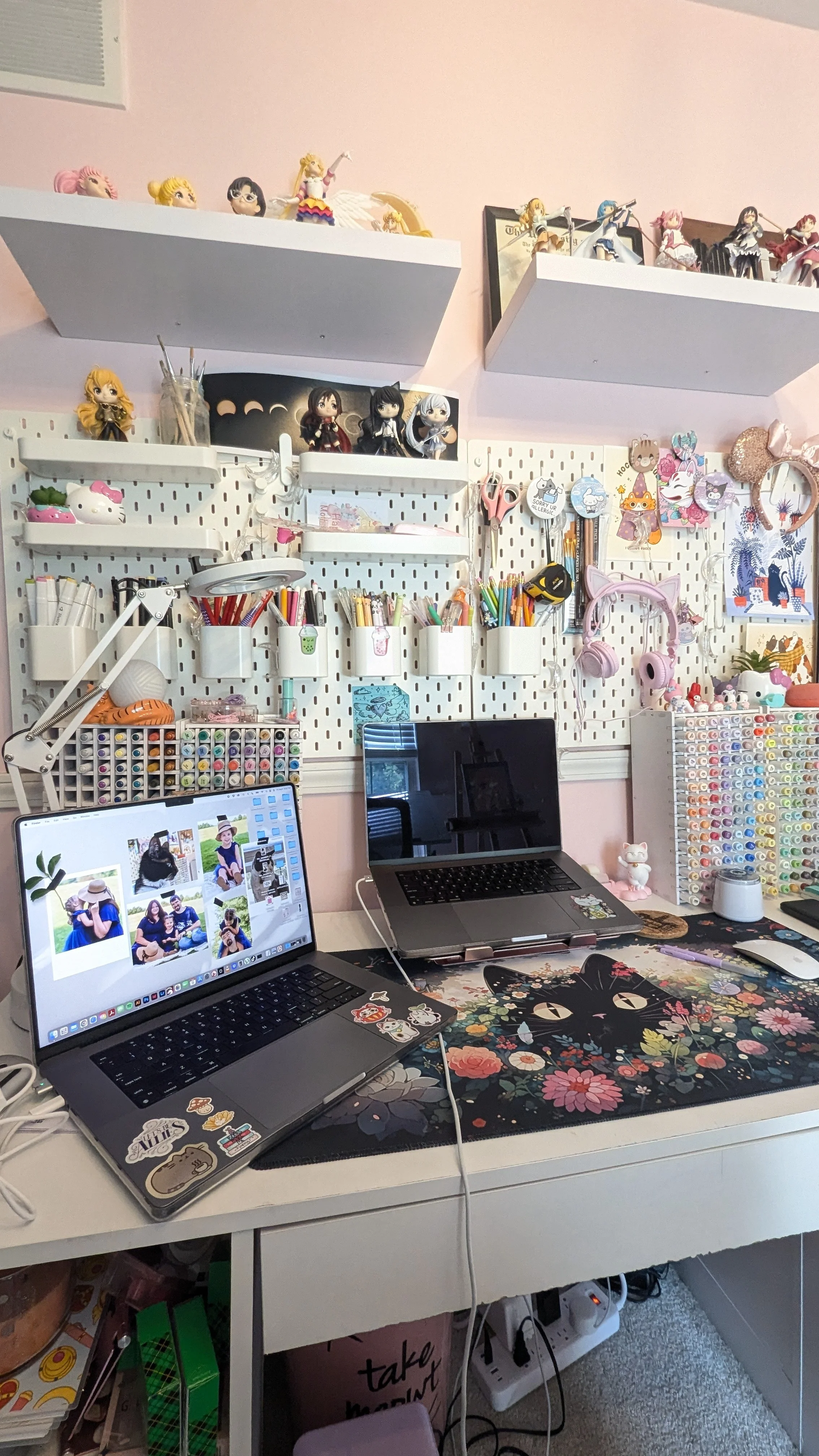 A workspace with two laptops on a desk, decorated with a colorful floral cat-themed mouse pad, surrounded by stationery, and a pegboard wall filled with organized pens, markers, and craft supplies. The wall display includes shelves with anime figurines, plush toys, and decorative items, creating a personalized creative area.