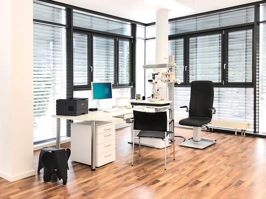 Augenarztpraxis mit Untersuchungstisch, Mikroskop, Computer und Stühlen in einem modern eingerichteten Raum mit Holzfußboden und großen Fenstern mit Rollos.