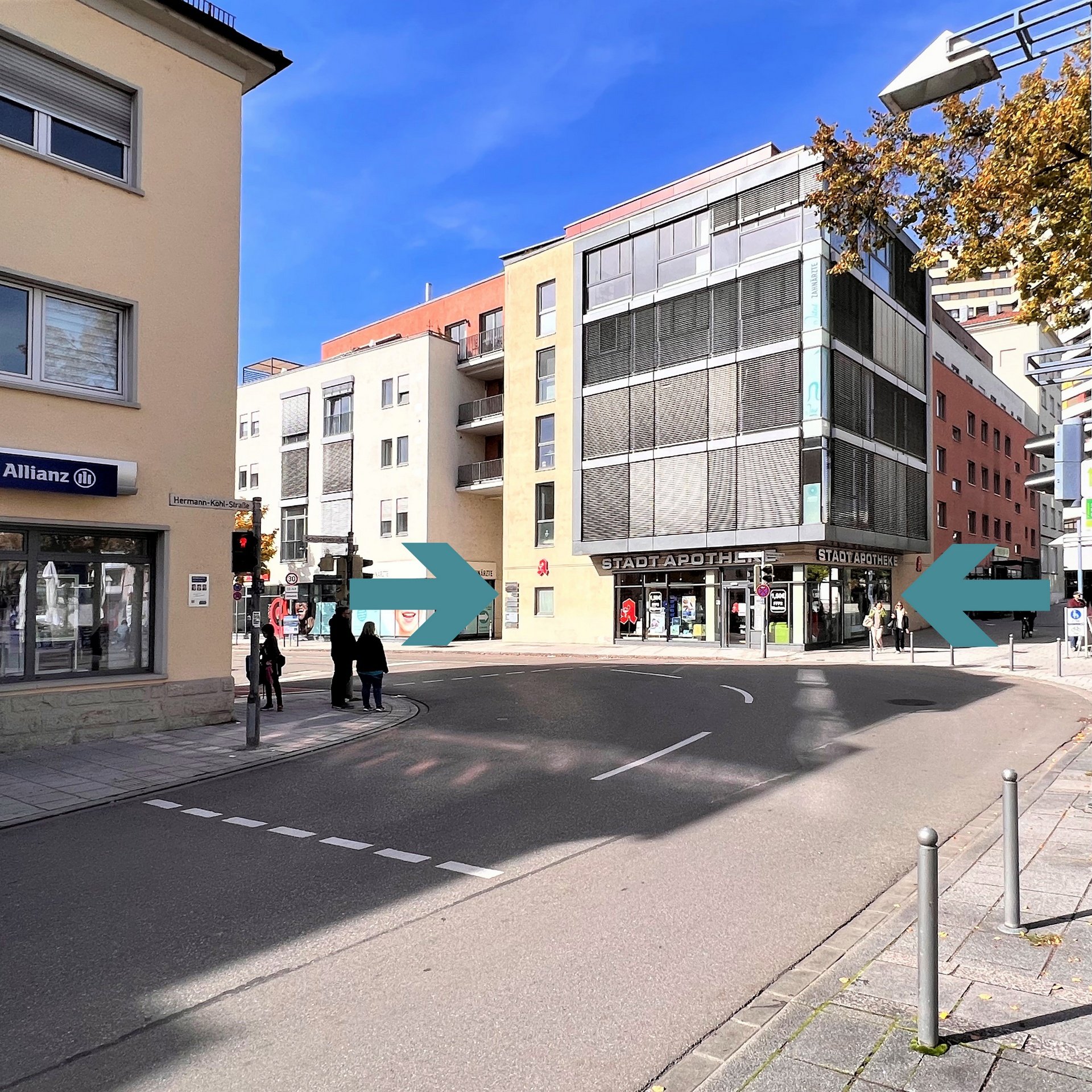 Straßenszene mit Fußgängern an einer Ecke, modernem Krankenhaus in der Mitte, im Hintergrund blaue Himmel, gelbe und graue Gebäude, Straßenlaternen und Verkehrsschilder.