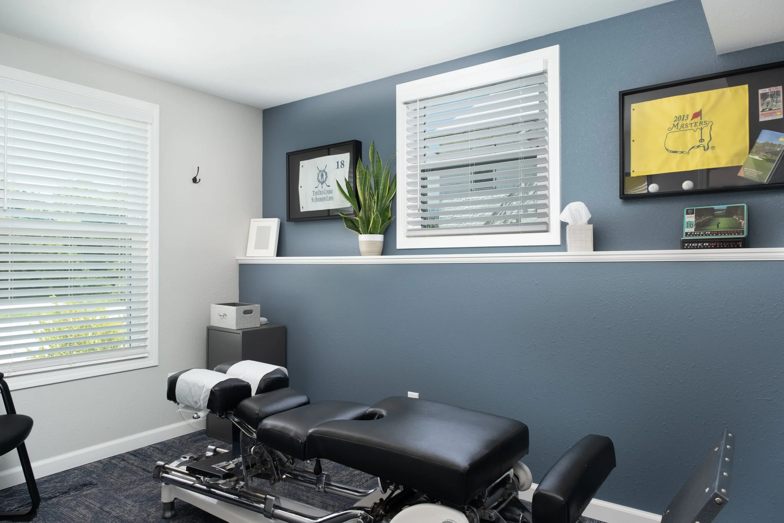 A chiropractic or medical examination room with a black adjustable treatment bed, a black chair, and window blinds, decorated with framed pictures, a potted plant, a box of tissues, and sports memorabilia on the walls.